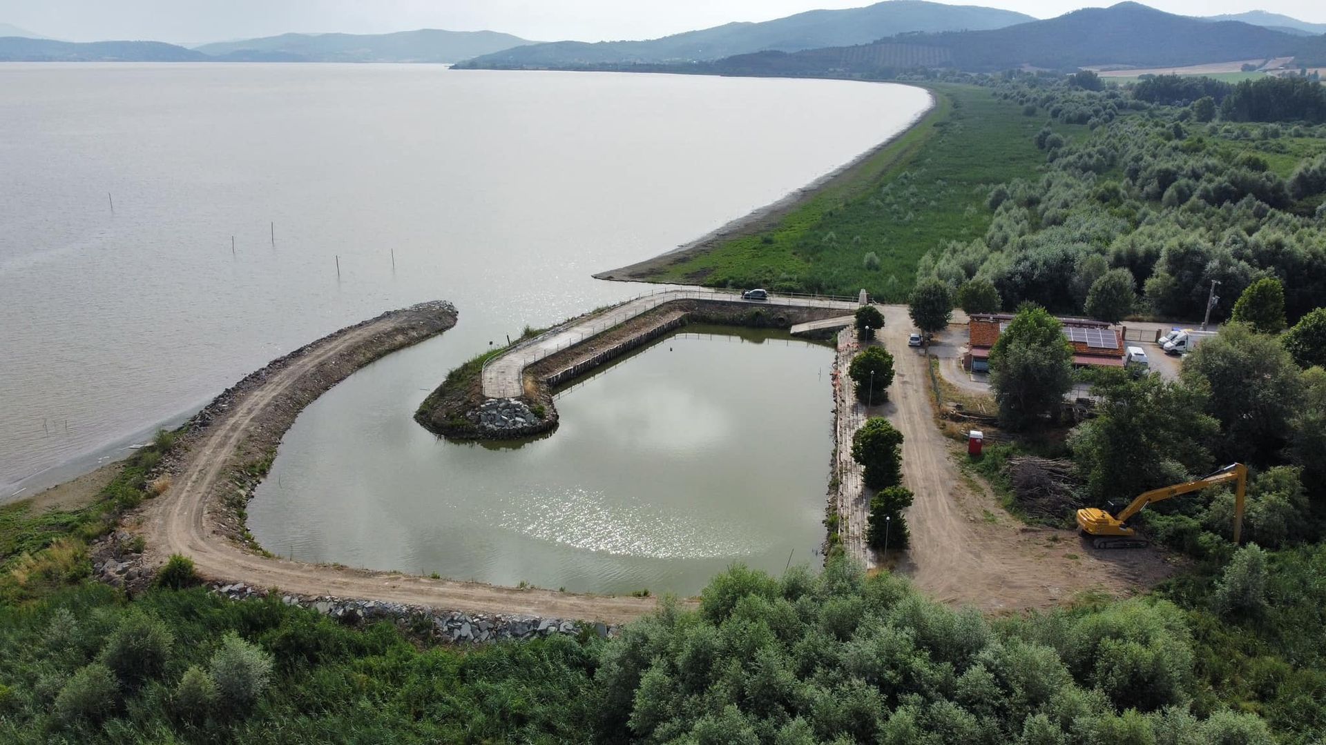 Vista aerea di un porto artificiale su un lago con una diga frangiflutti curva, circondato da alberi e montagne sullo sfondo.