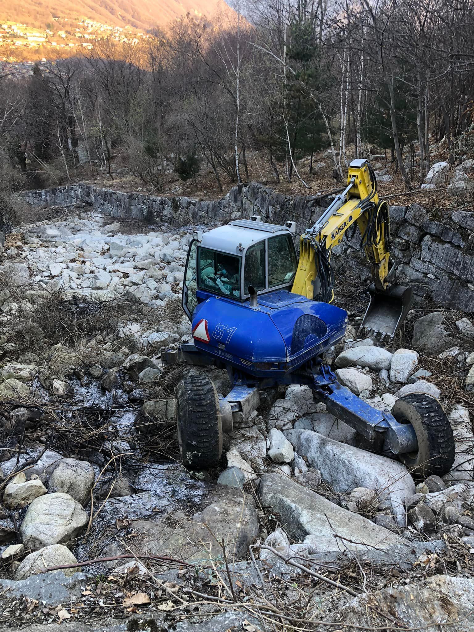 Escavatore blu e giallo in una zona rocciosa e boscosa.
