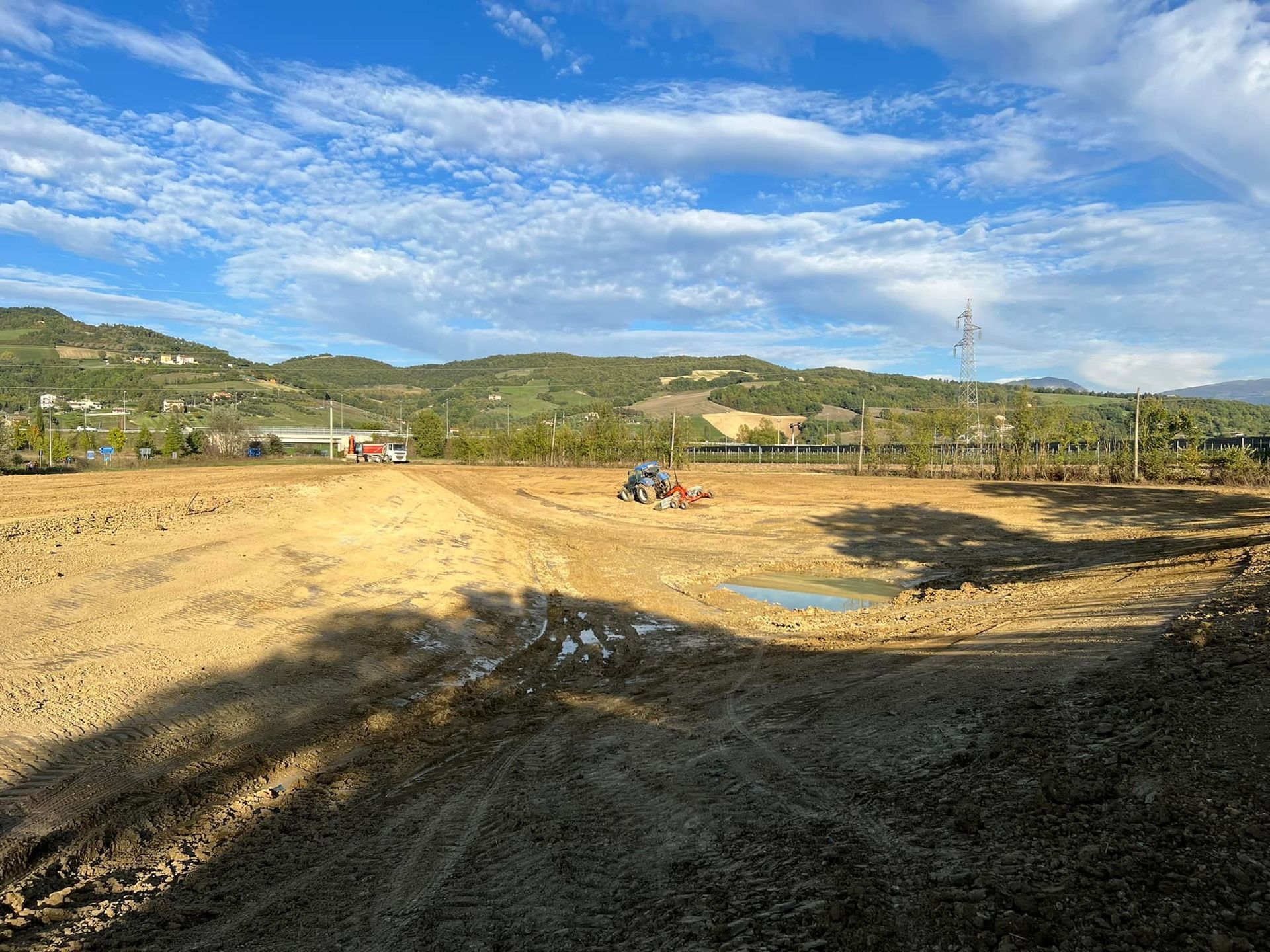 Un trattore che ara un campo marrone sotto un cielo azzurro brillante con nuvole sparse e uno sfondo di colline.