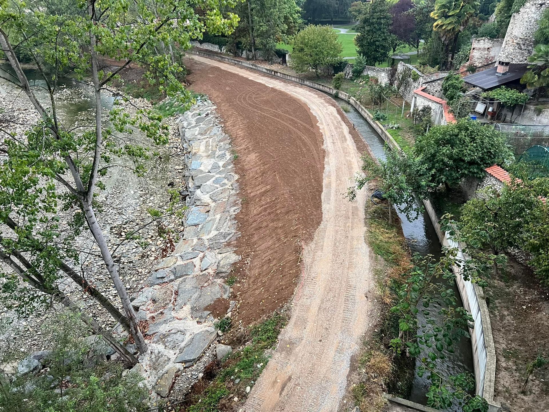 Sentiero sterrato che si snoda tra un muro di pietra e un canale d'acqua con alberi.