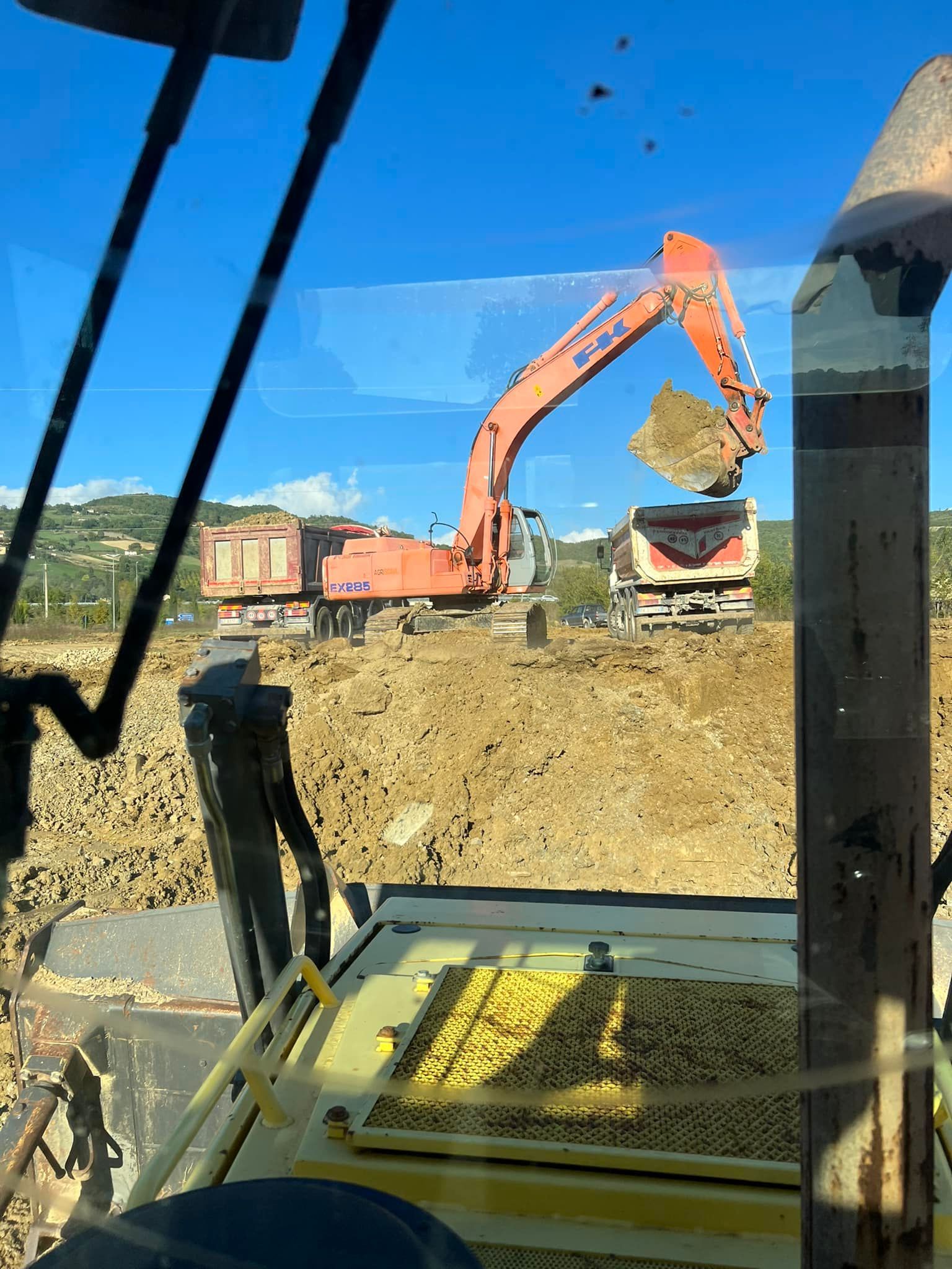 Un escavatore carica un camion con cassone ribaltabile pieno di terra in un cantiere edile sotto un cielo azzurro.