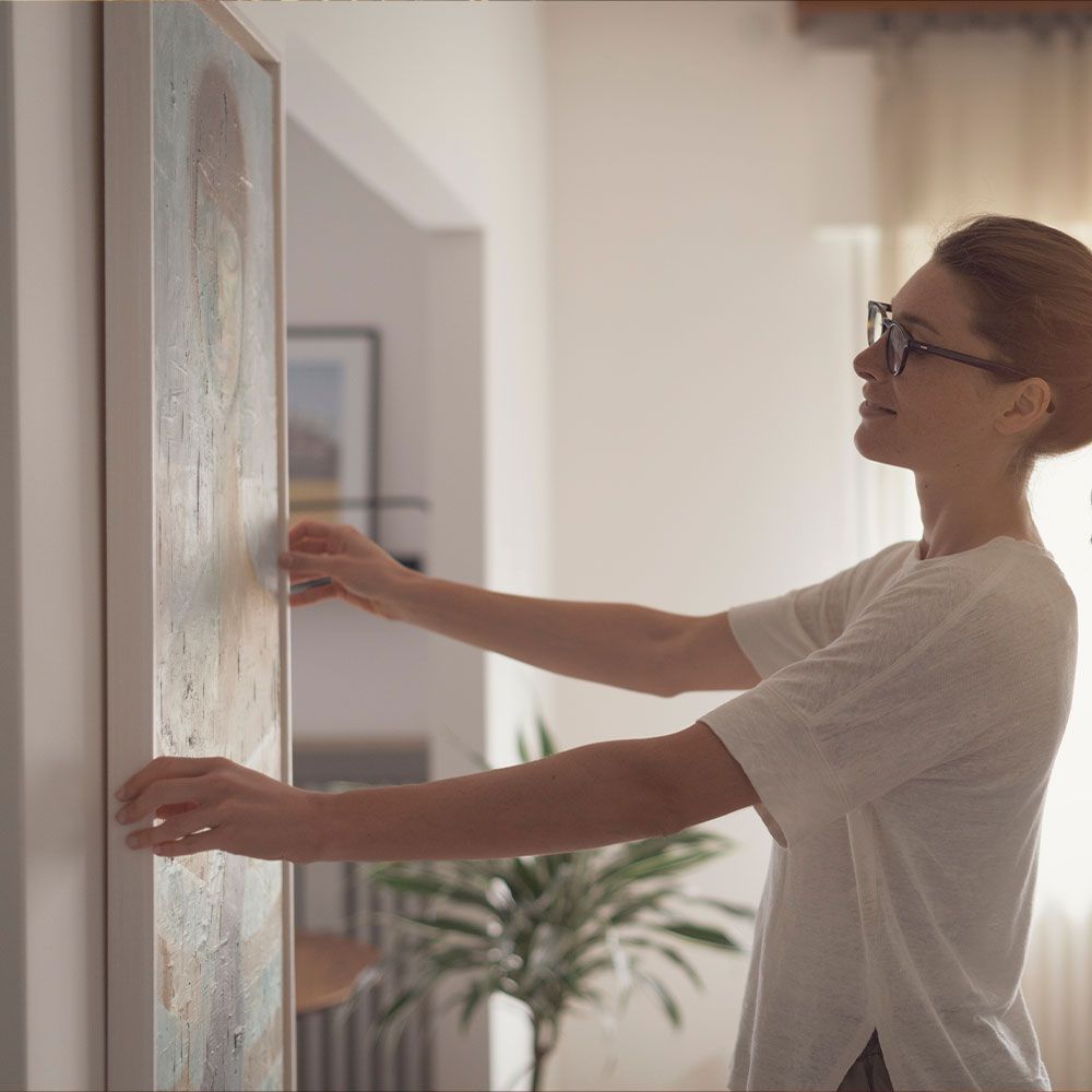 A woman wearing glasses is hanging a picture on a wall