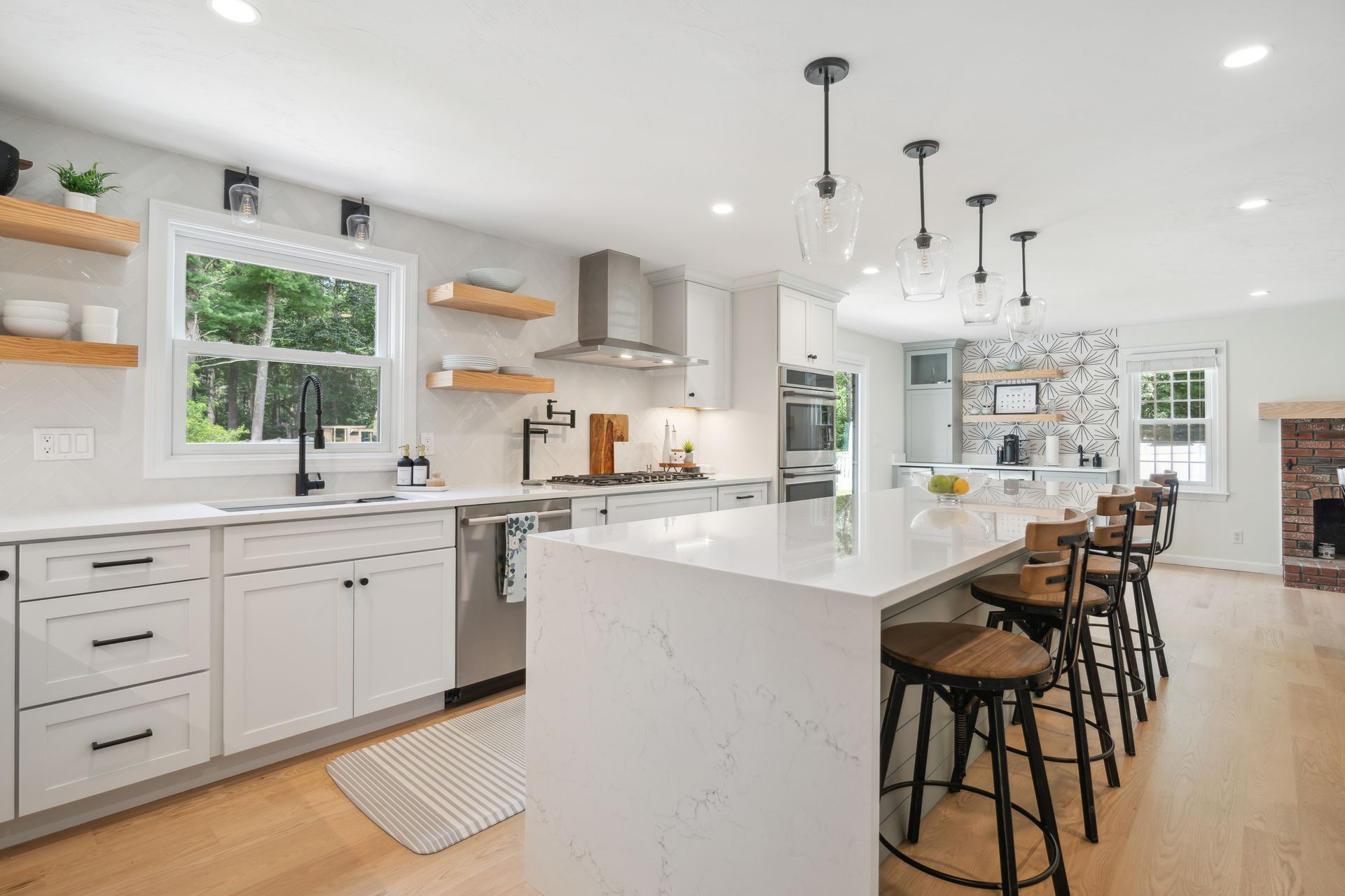 Modern white kitchen with island, wooden shelves, and bar stools. Bright with natural light.