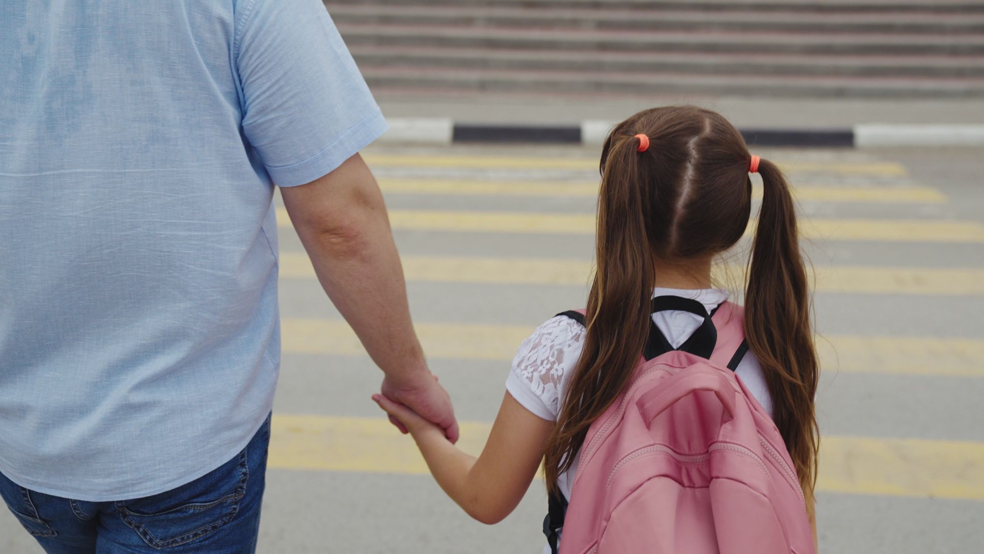 A person holding a child's hand walks across a crosswalk. The child wears a pink backpack.