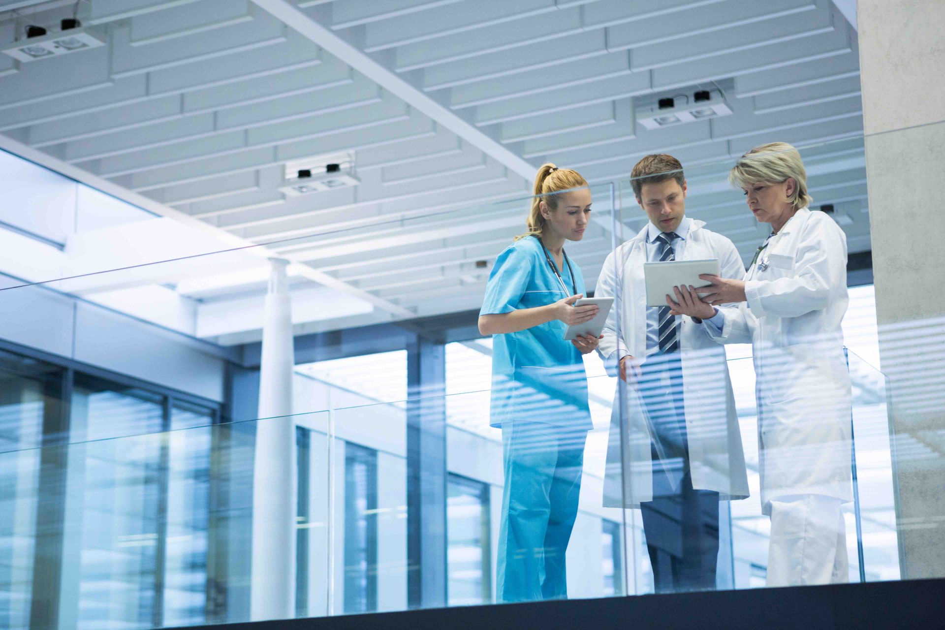Three medical professionals in a modern hallway reviewing documents on tablets, one in scrubs, two in lab coats.