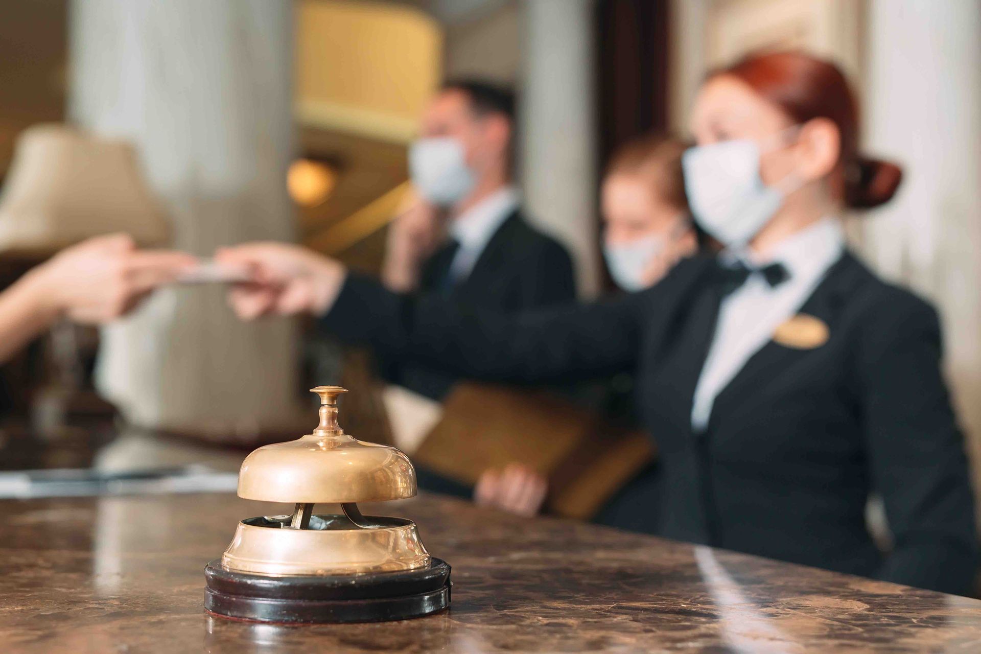 Hotel reception desk with bell; staff wearing masks assists a guest.