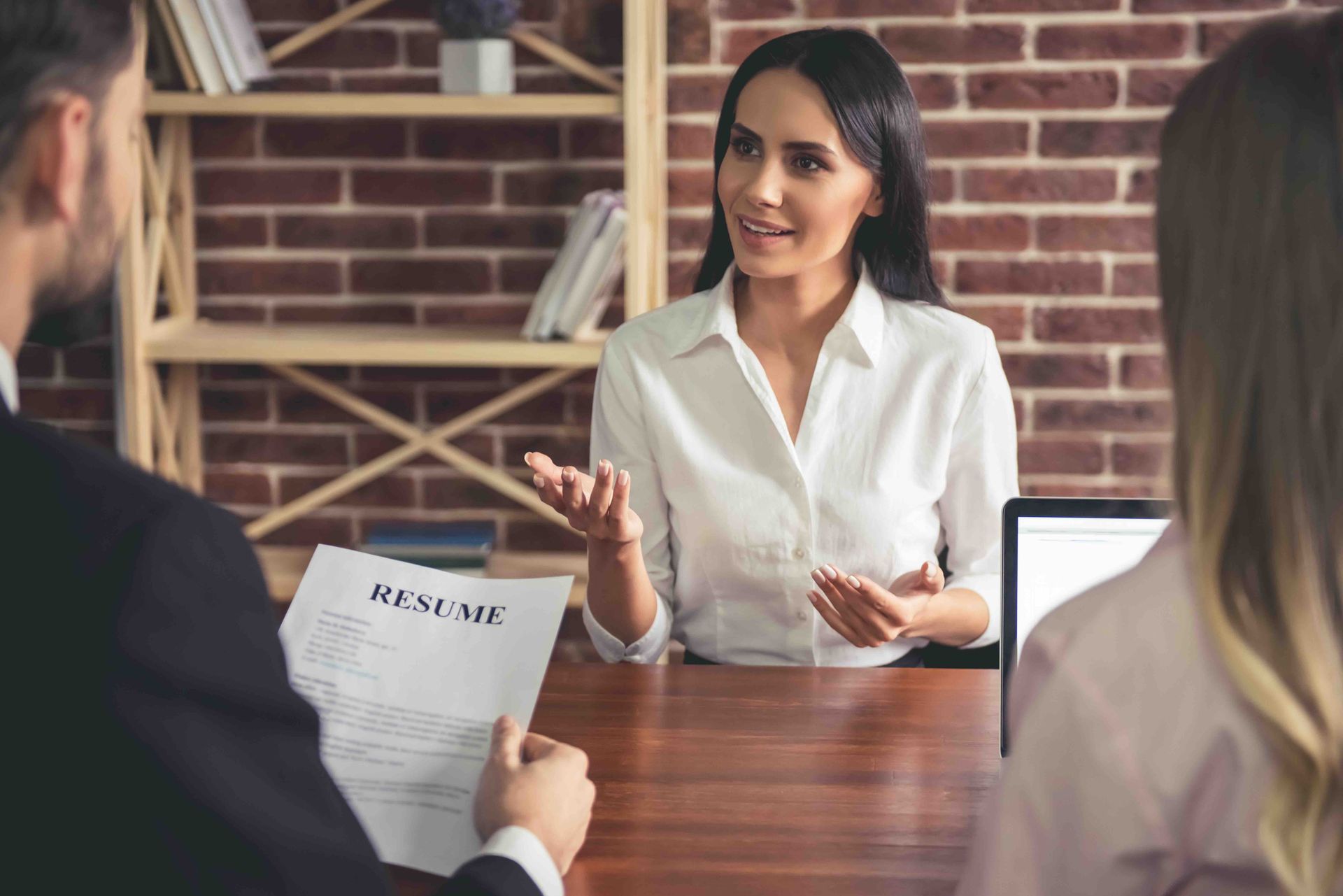 Woman in white shirt gestures, talking to two people seated across a desk. A resume is visible.