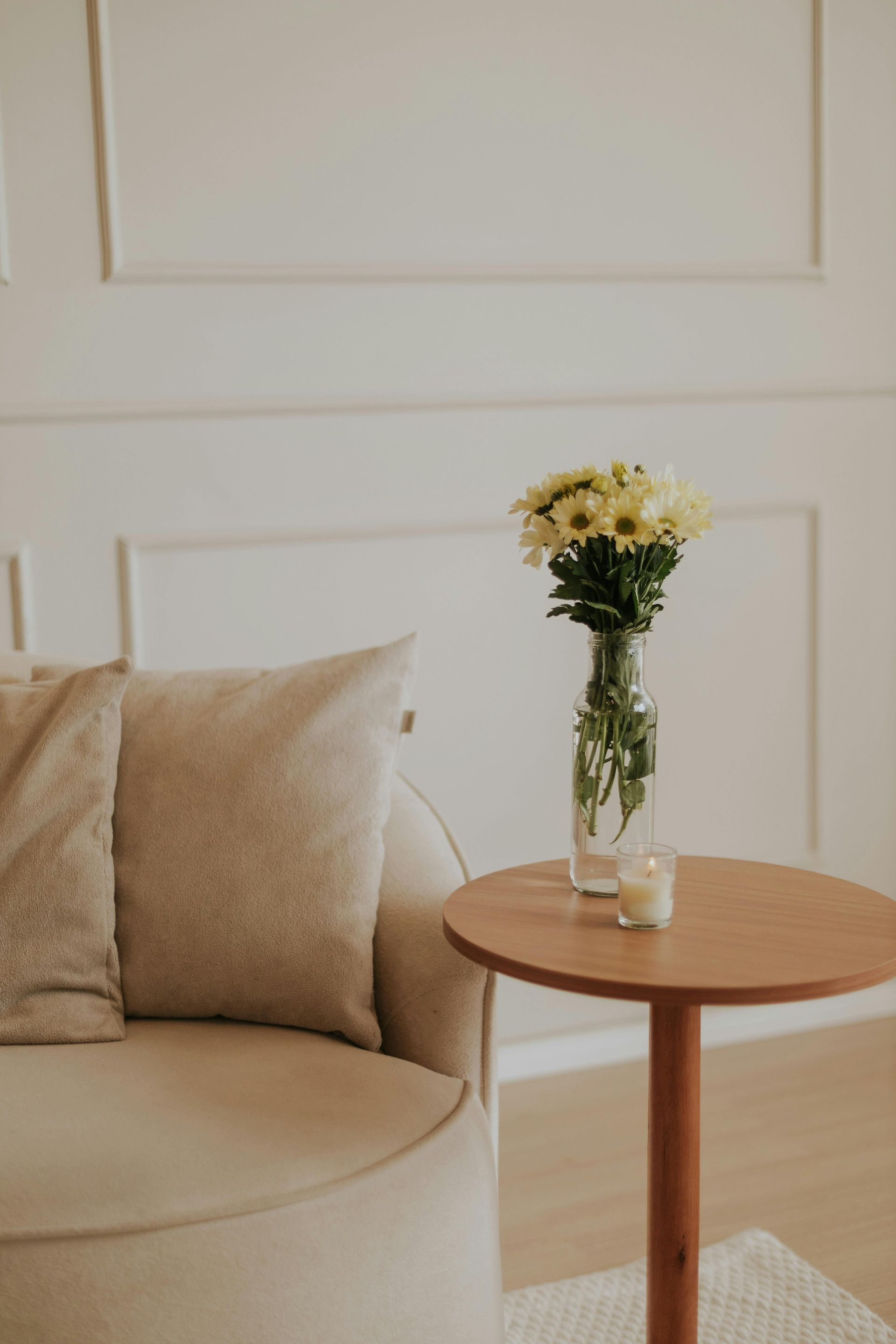 A beige armchair and round wooden table with yellow flowers in a vase and a candle against a white wall.