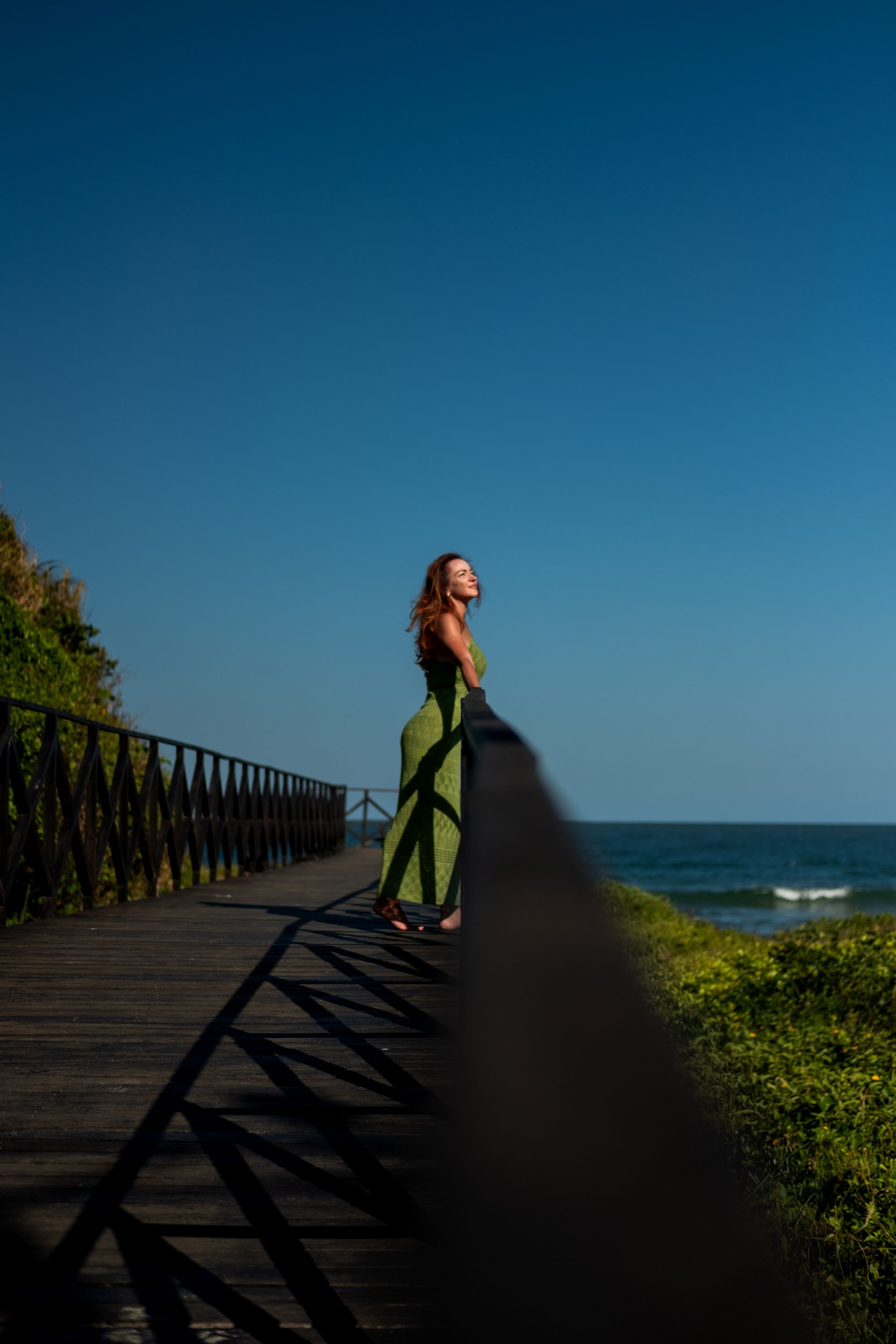 Mulher de vestido verde, em um calçadão de madeira com vista para o oceano, sob um céu azul.
