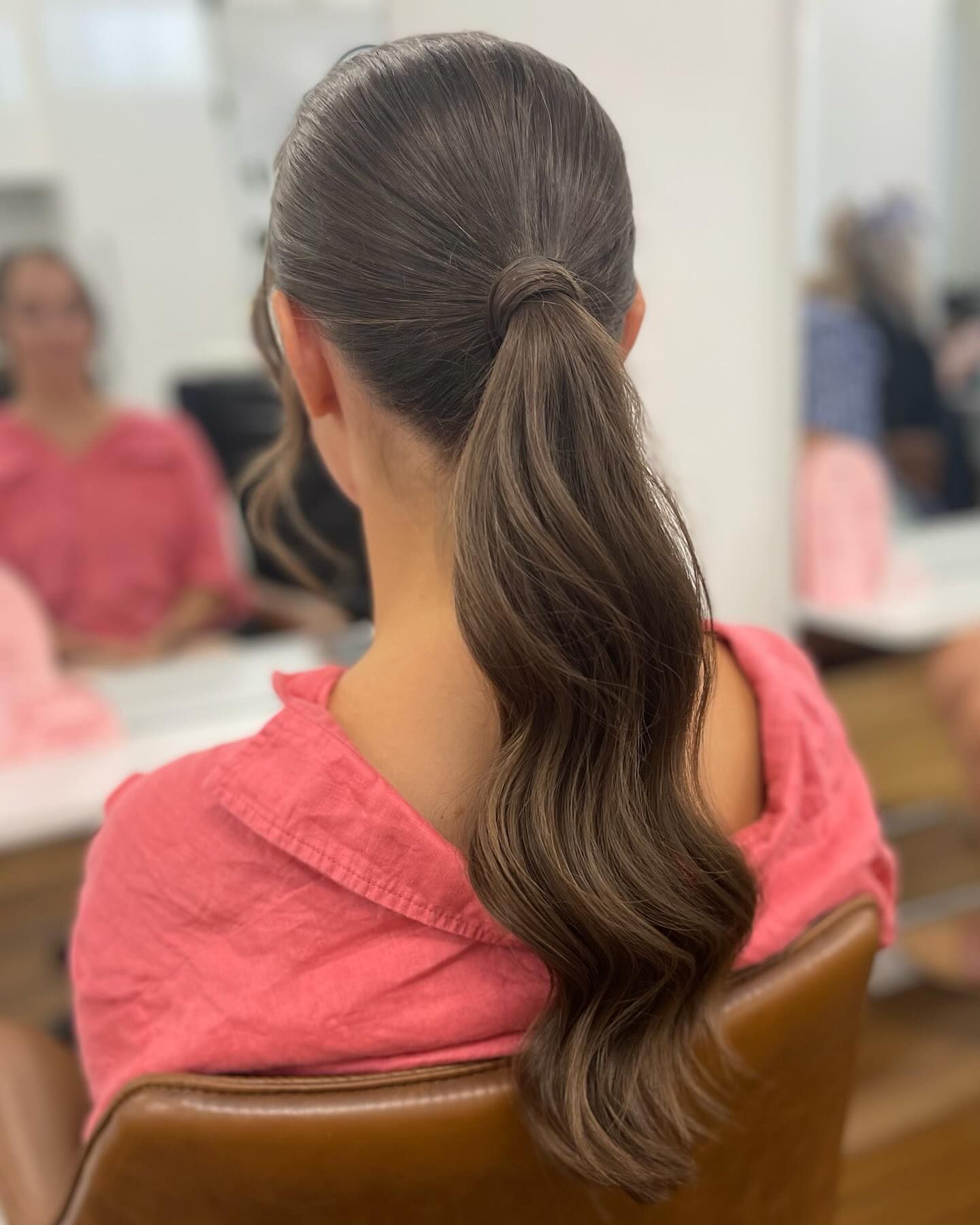 Person with a long wavy ponytail wearing a pink top, seated in a salon chair and viewed from behind — Pose Hair Design in Orange, NSW
