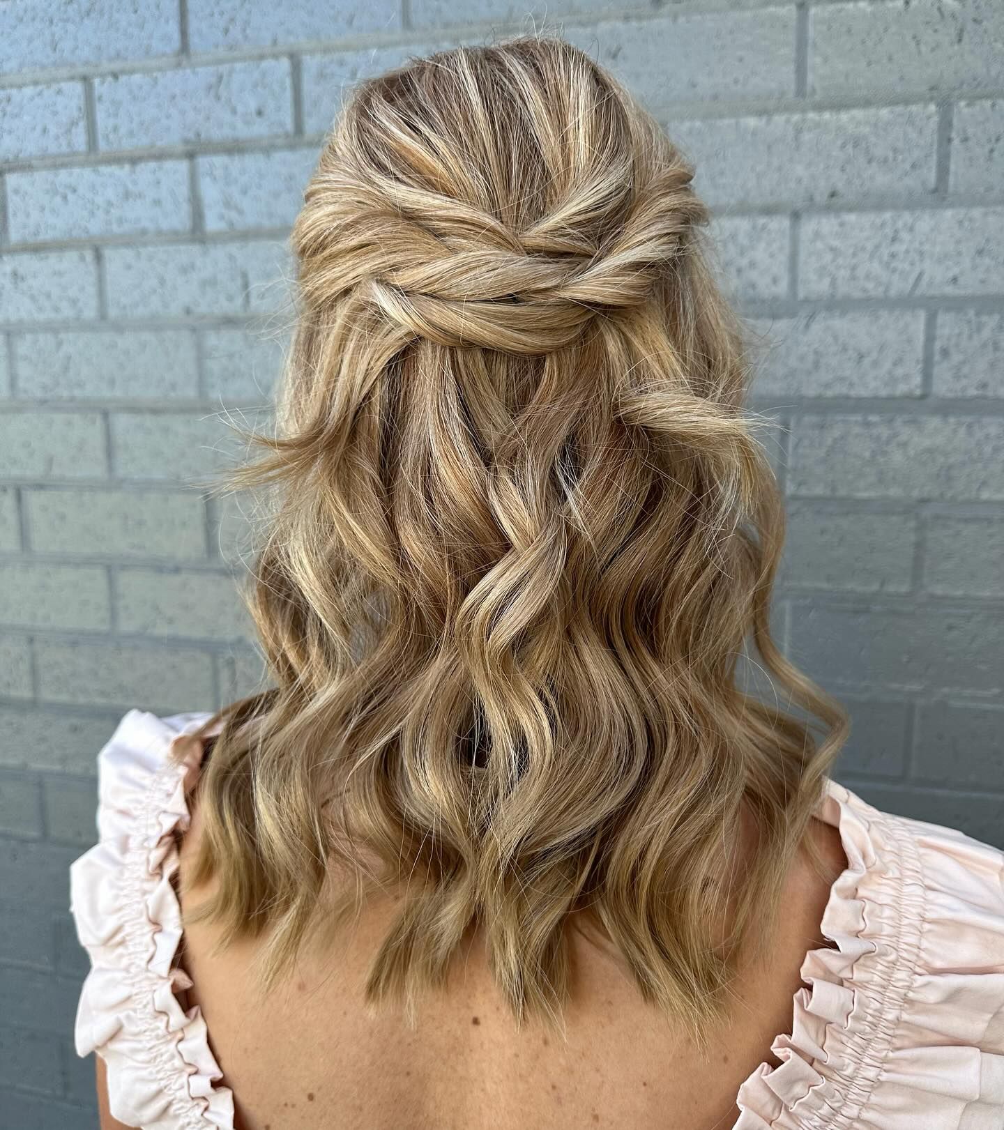 Back view of wavy blonde half-up hairstyle against a grey brick wall — Pose Hair Design in Orange, NSW