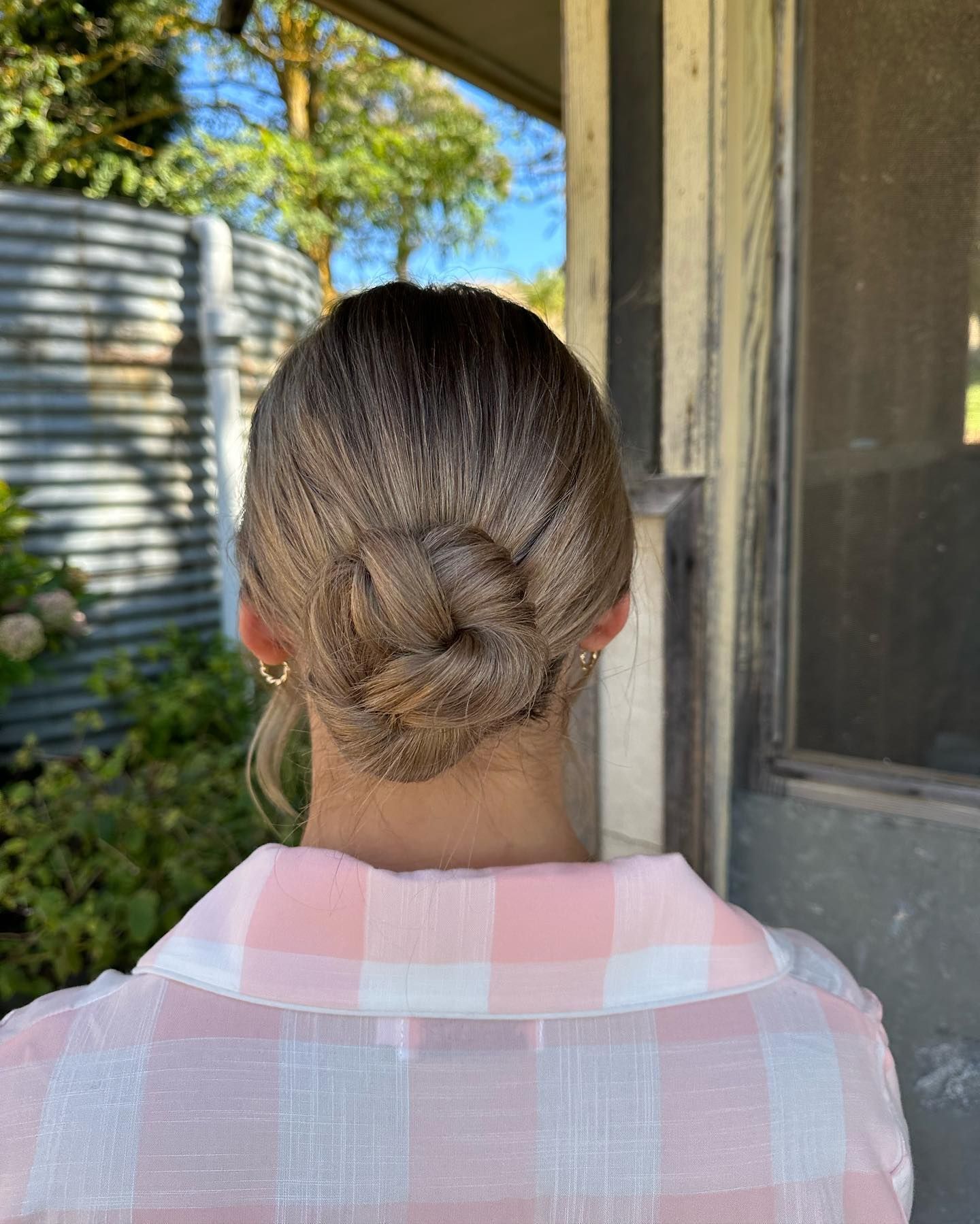 Back view of a person with a twisted bun hairstyle wearing a pink plaid shirt outdoors by a doorway — Pose Hair Design in Orange, NSW