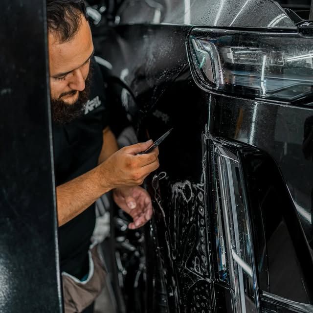 Man applying film to black car bumper, using a small tool. Shop setting.