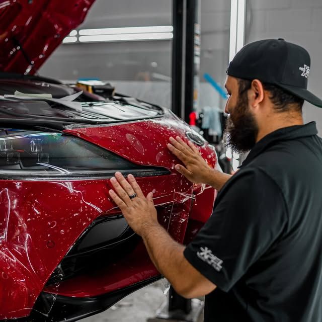 Man applying protective film to a red car bumper in a workshop.