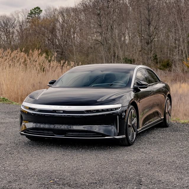 Black Lucid Air electric sedan parked on gravel road with tall grass and trees in background.