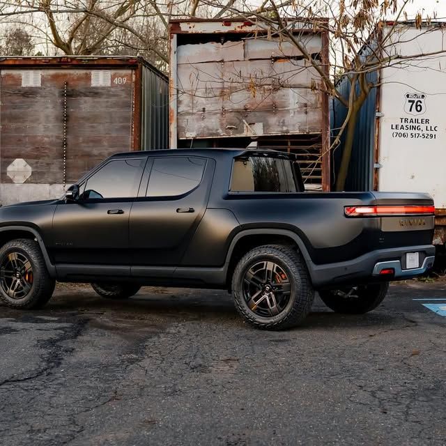 Black Rivian electric pickup truck parked outdoors, near shipping containers.