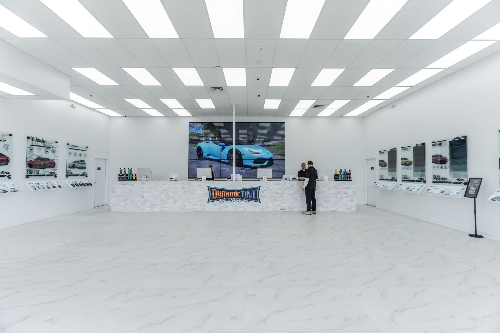 Bright, modern retail space. A person stands at a counter in front of a large screen. Walls have display cases with product images.