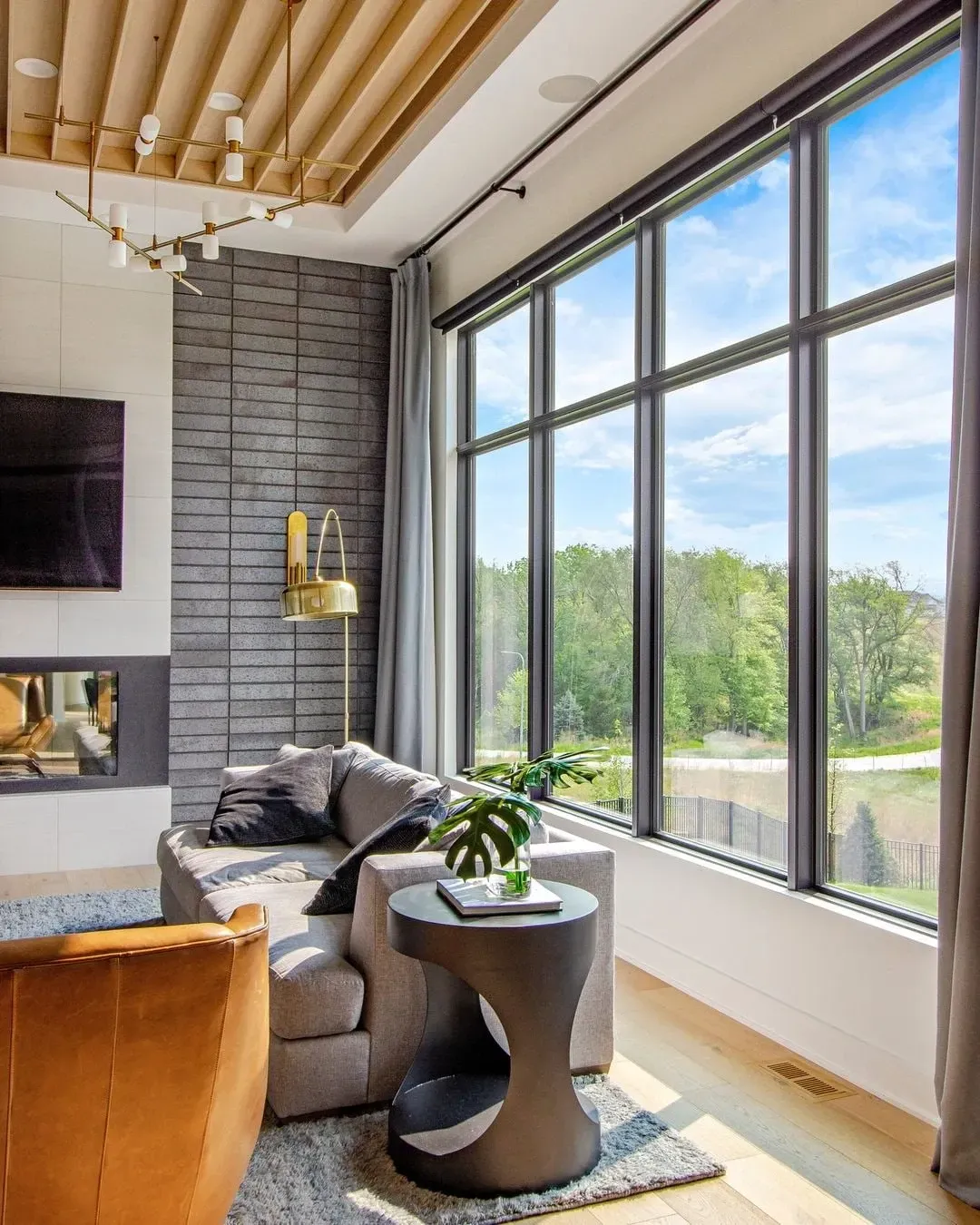 Living room with large windows overlooking a green landscape, gray sofa, fireplace, and modern light fixtures.