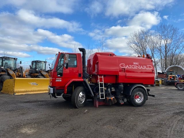 Camion balayeuse rouge sur un parking asphalté ; stationné à côté d'engins de chantier sous un ciel nuageux.