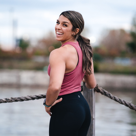 A woman in a pink tank top and black pants is standing next to a rope fence and smiling.