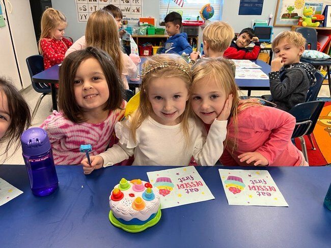 Three young girls smiling at a table with a toy cake and other children in a classroom.