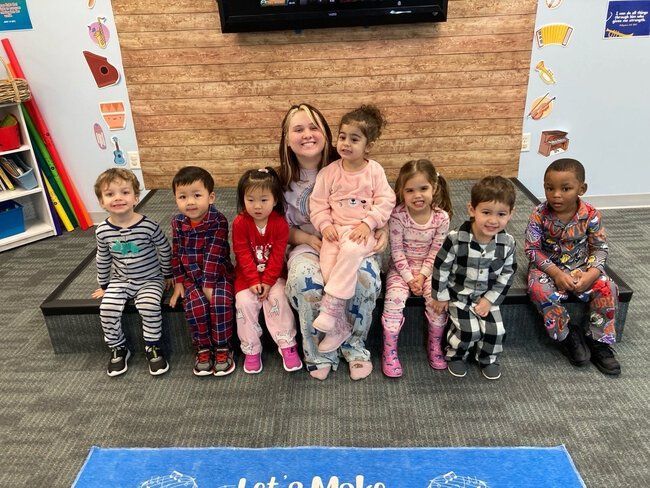 A teacher and children in pajamas sit on a stage indoors. They are smiling.