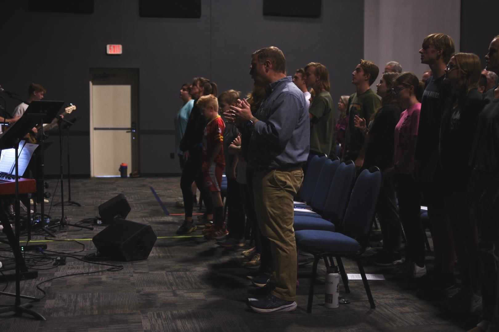 People singing and clapping in a dimly lit room, with a band on stage.