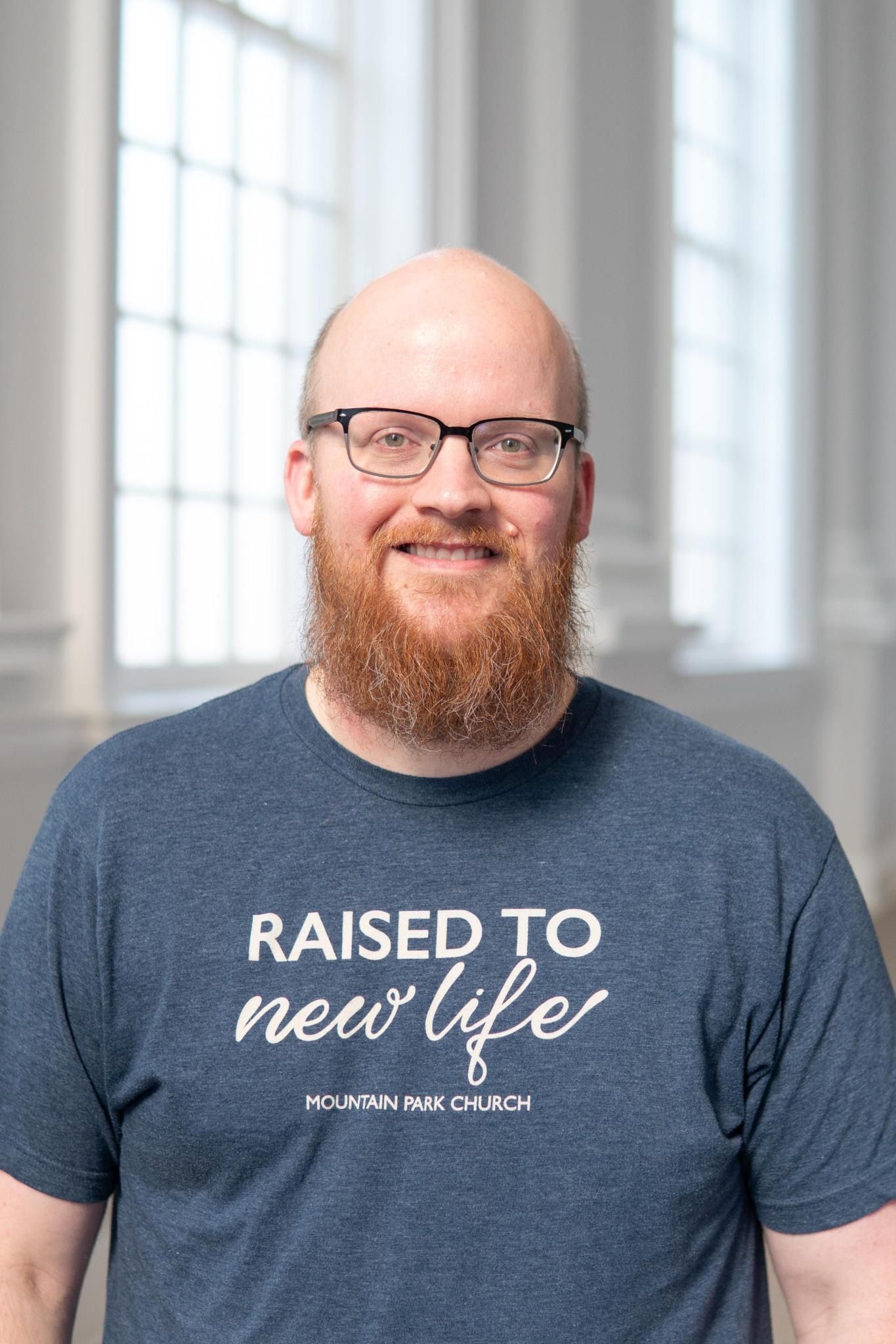 Man with a red beard and glasses smiles, wearing a gray shirt, neutral background.
