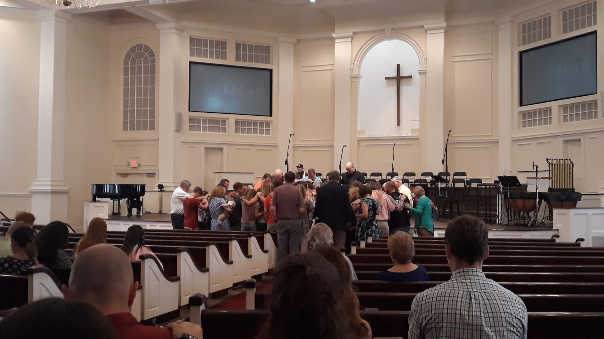 People singing in a church. Choir in center, cross on wall, screen and pews visible.