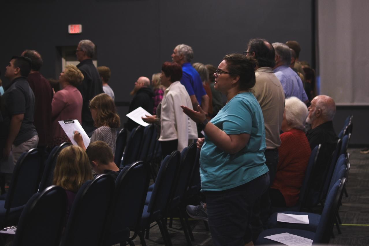 People in a large room, some singing and raising hands. Dark chairs, a woman in teal stands out.