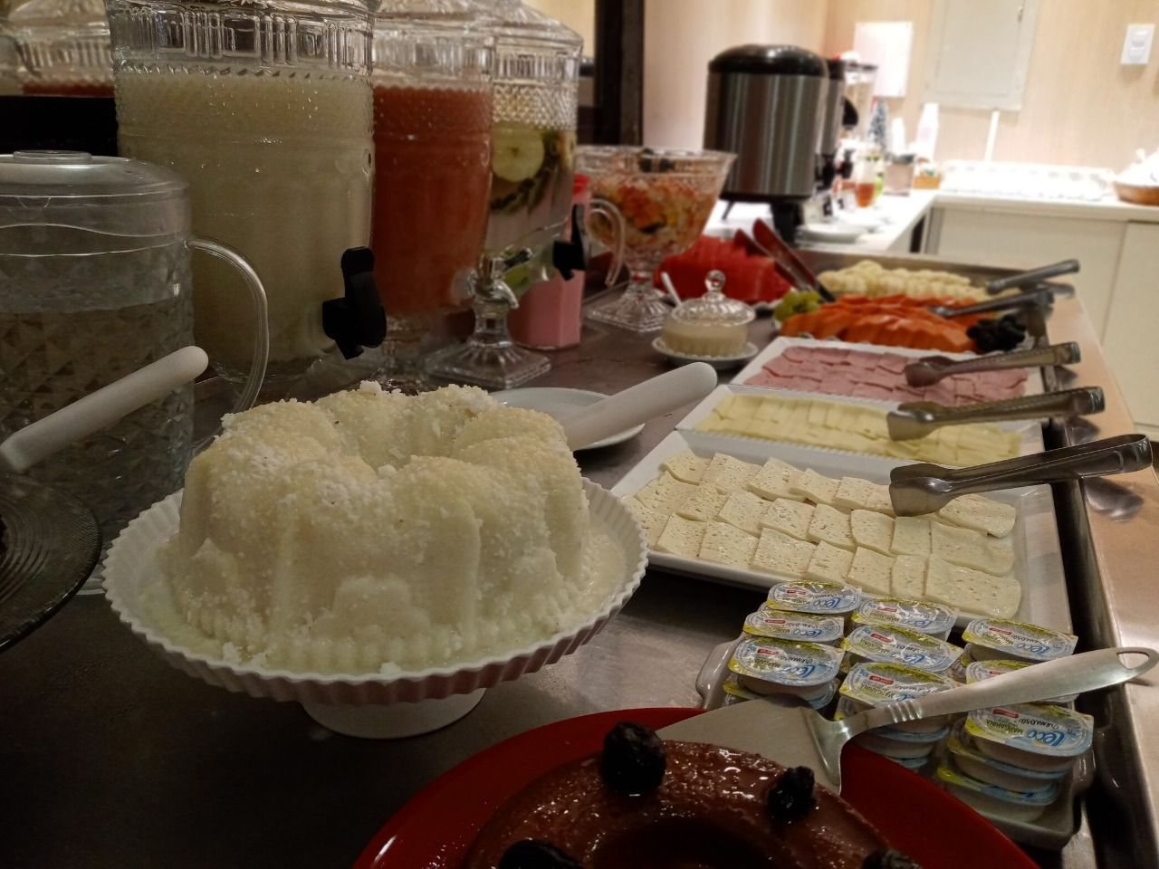 Buffet de café da manhã com bolos, sucos, queijos e frios fatiados em um balcão.