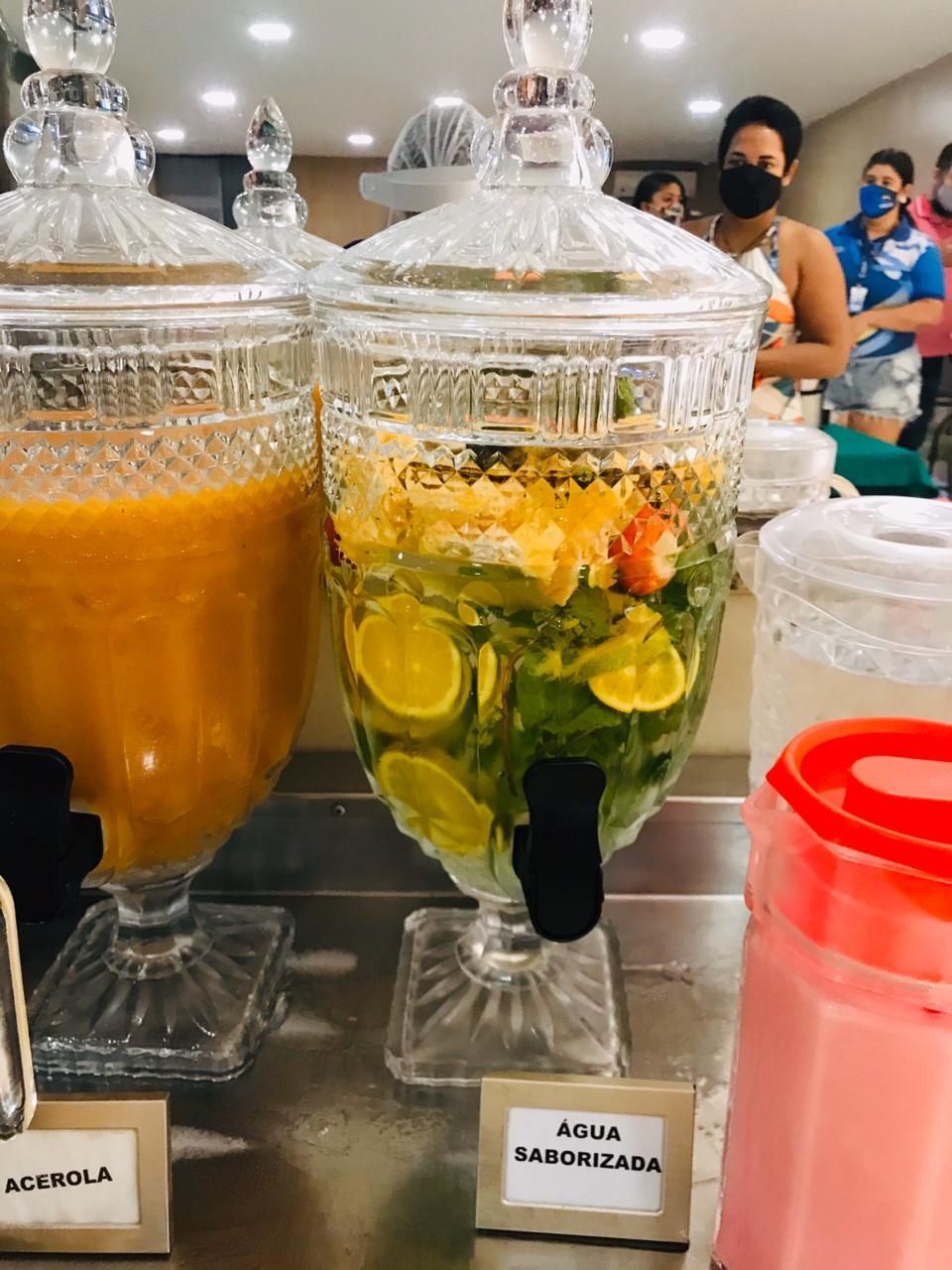 Grandes dispensadores de vidro com água aromatizada com frutas e suco em um bufê. Pessoas ao fundo.