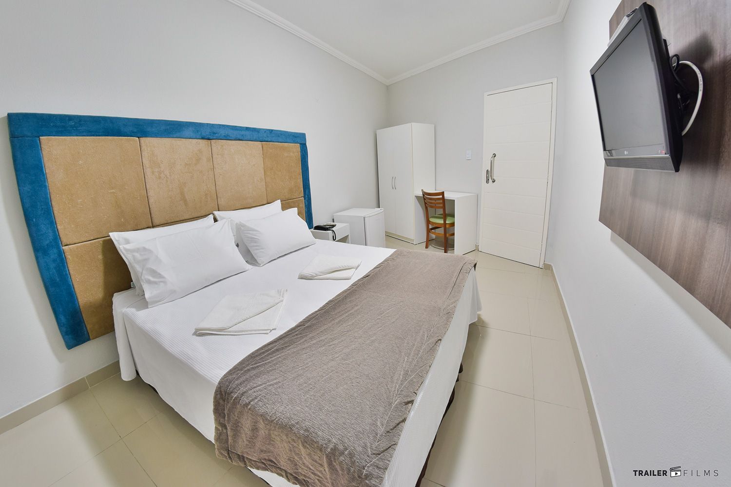 Quarto de hotel com cama, TV e armário; paredes brancas, tons neutros.