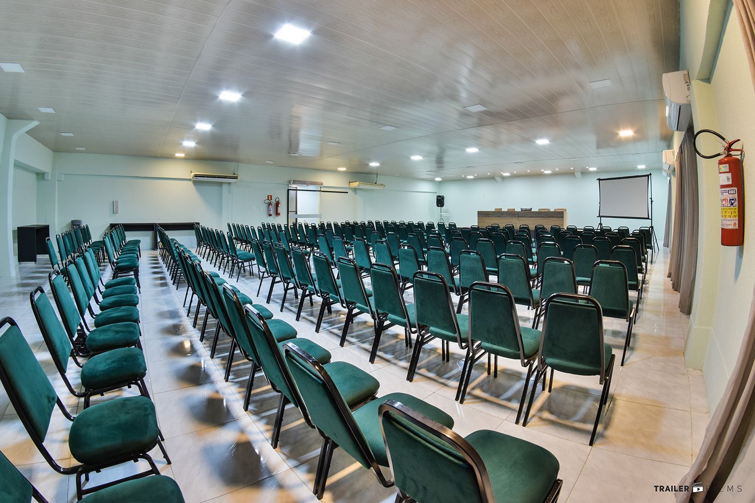 Auditório vazio com fileiras de cadeiras verdes voltadas para um palco com uma tela e uma mesa.