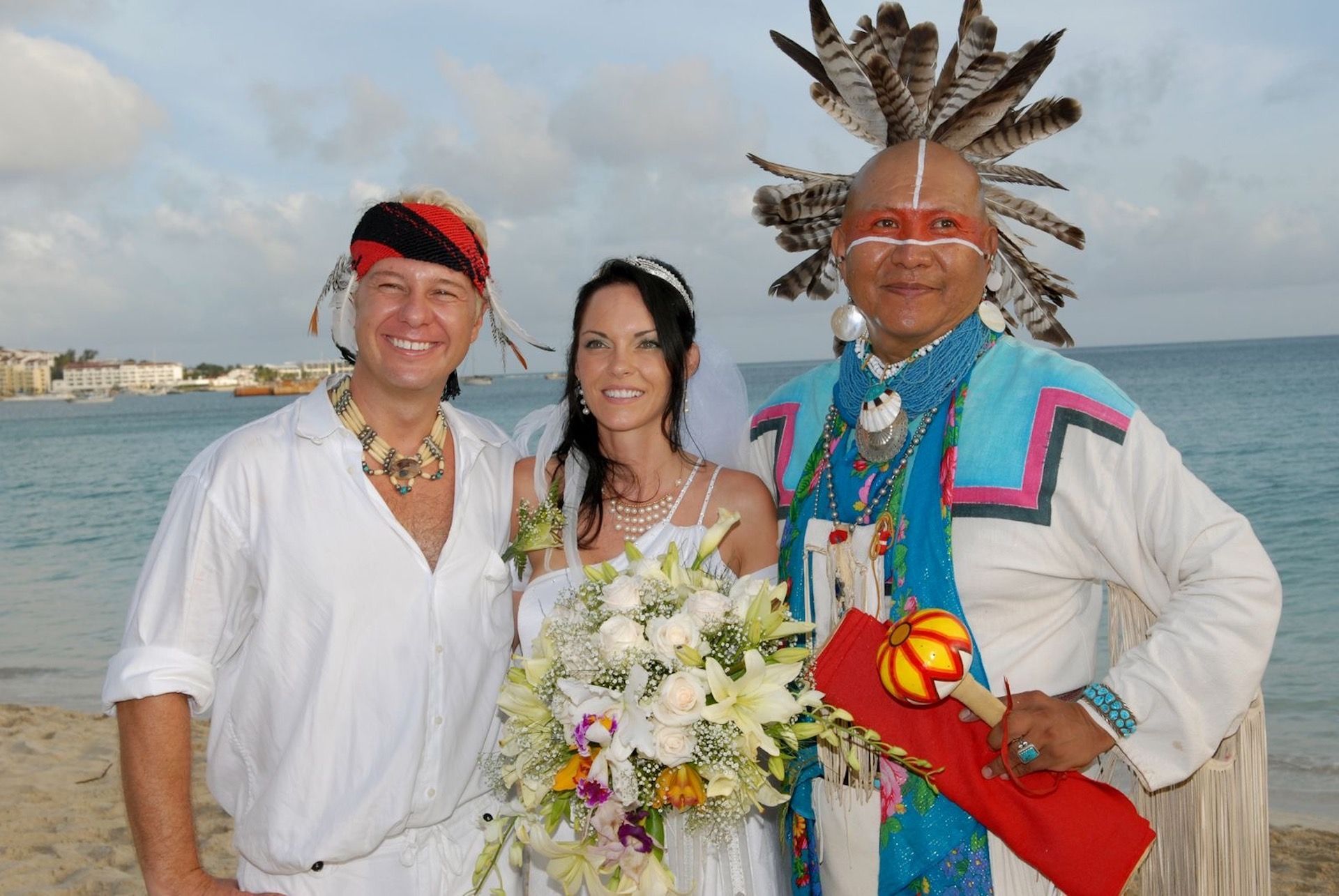 Bride and groom with Native American officiant on a beach, smiling. Bright clothing, feathers, flowers, and ocean background.