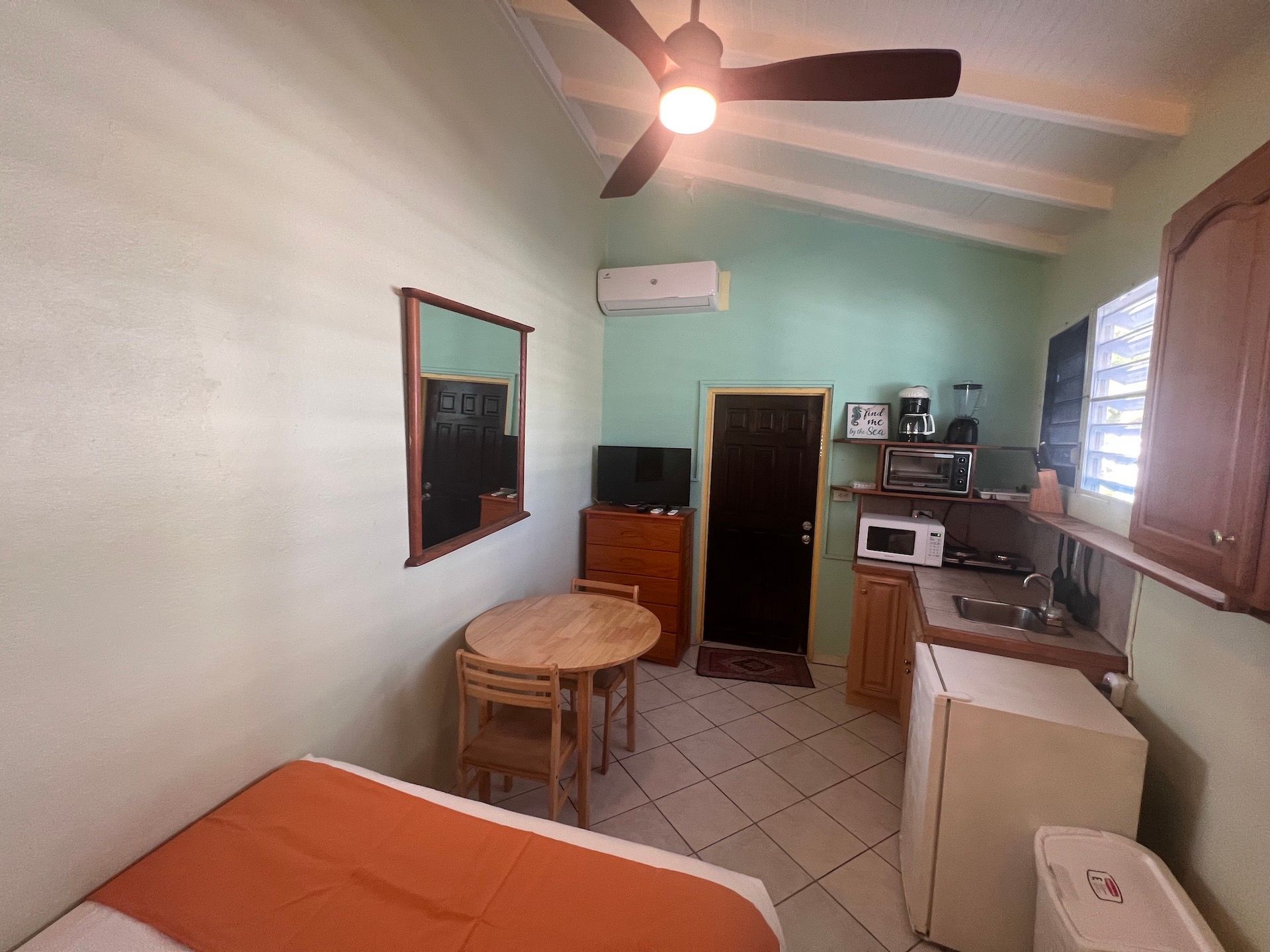 Small, studio apartment with bed, table, kitchenette, and dark brown door; light green walls.