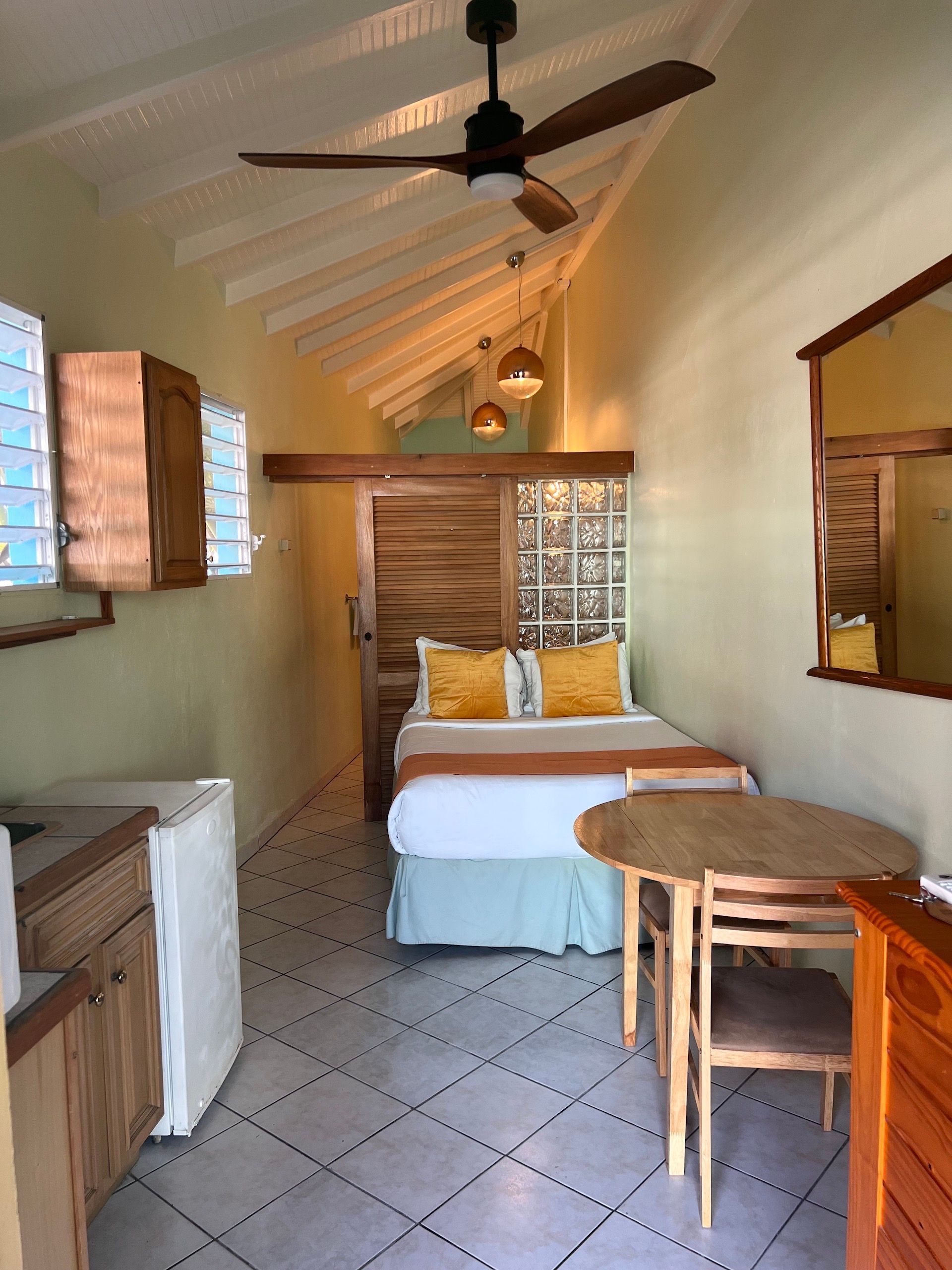 Cozy studio apartment with bed, small dining table, kitchenette, and vaulted ceiling; light green walls, tile floor.