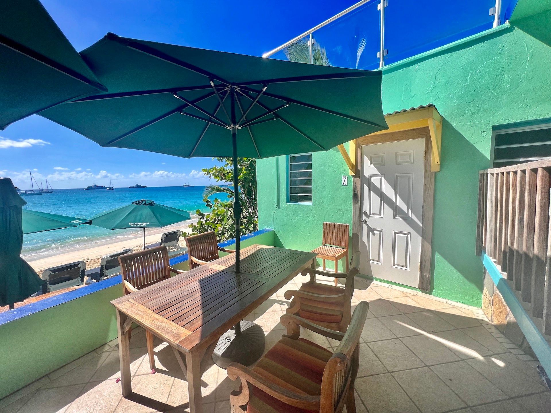 Outdoor patio with table, chairs, and green umbrella, overlooking a beach with turquoise water.