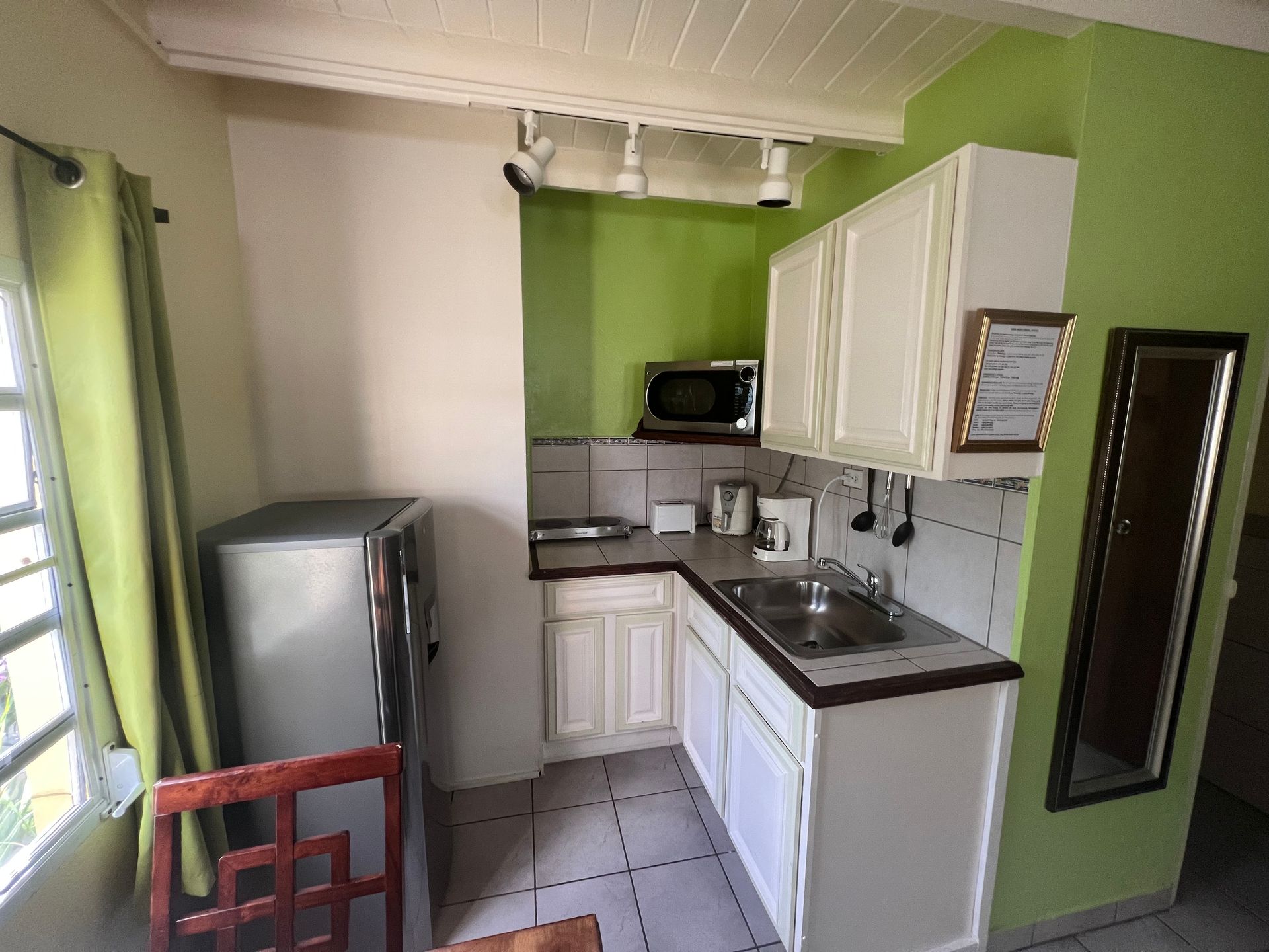 Small kitchen with white cabinets, green wall, microwave, fridge, sink, and a window.