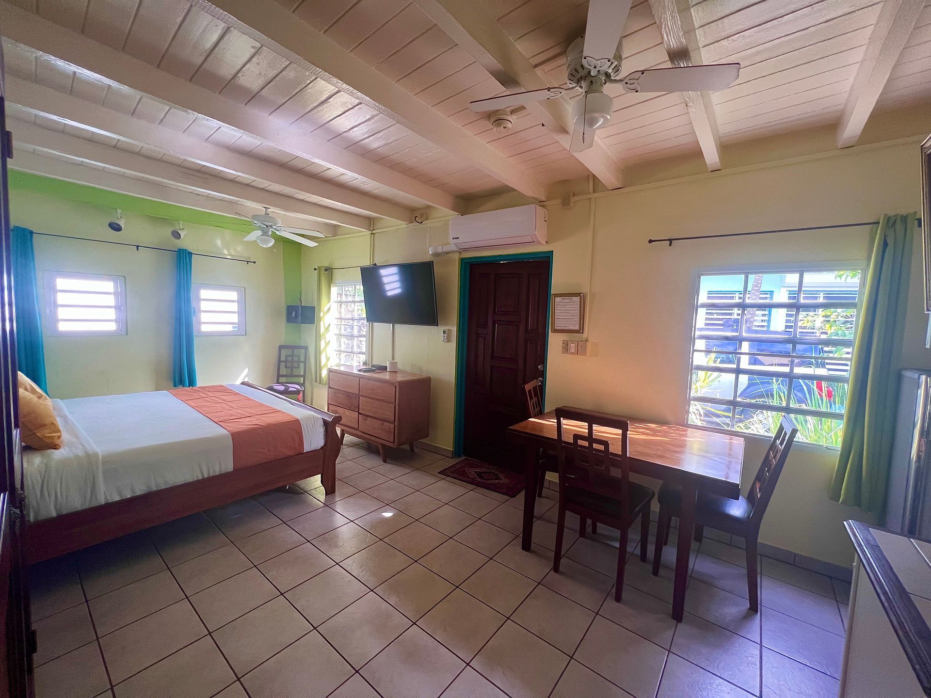 Bright studio apartment with bed, table, and dresser; has a window, door, and ceiling fan.