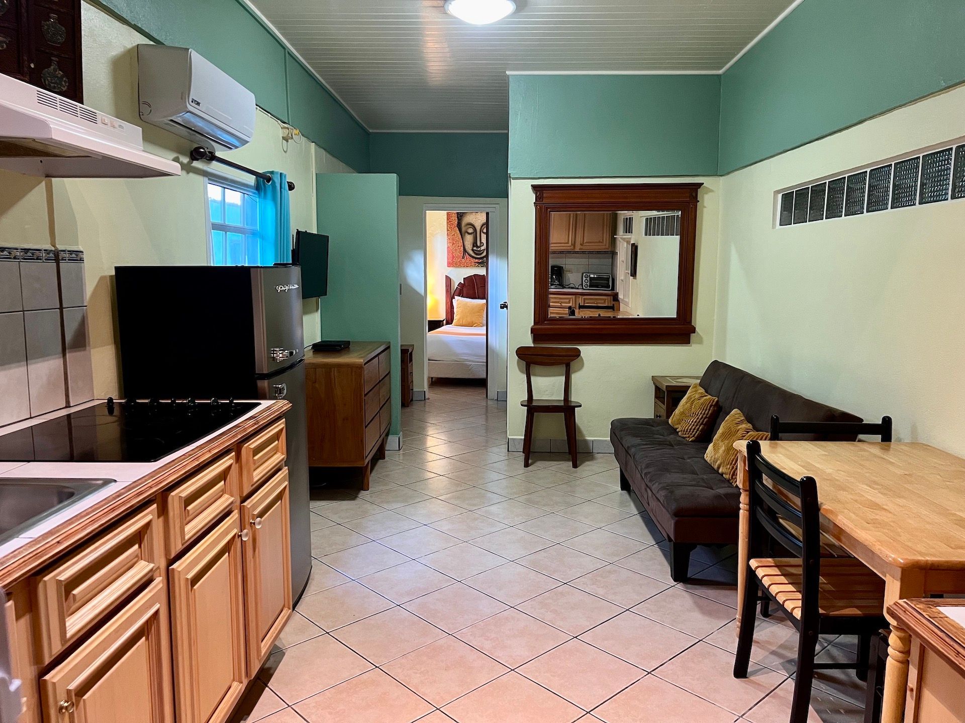 Small apartment interior with kitchen, living area, and a glimpse of the bedroom. Beige and teal walls, wood furniture.