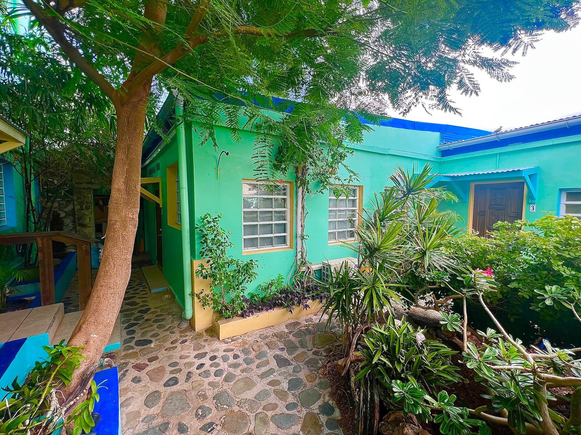 Green and blue Caribbean cottage with stone path, surrounded by lush greenery.