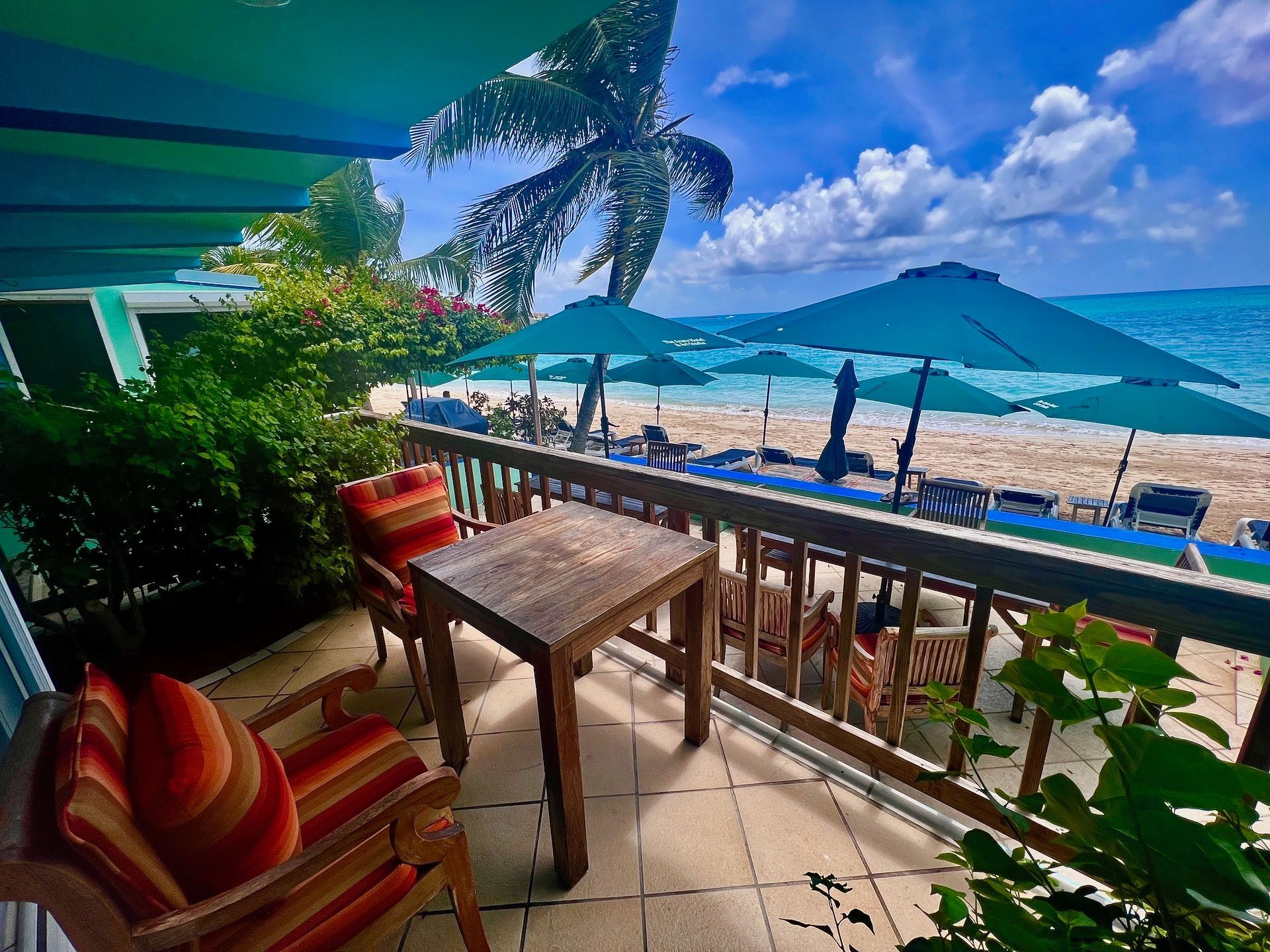 Balcony overlooking a beach with turquoise umbrellas, wooden furniture, and a palm tree.