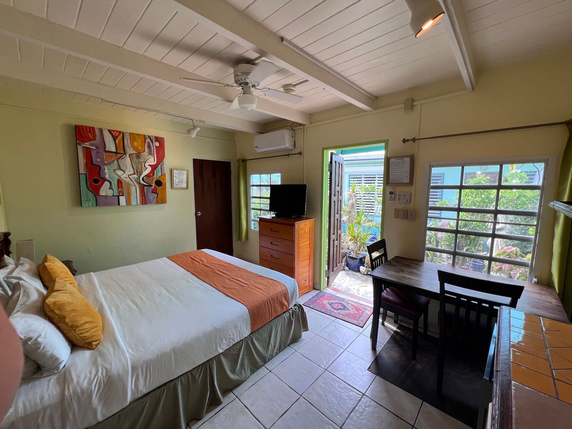 Bedroom with bed, art, dresser, table, open door to patio, and windows.
