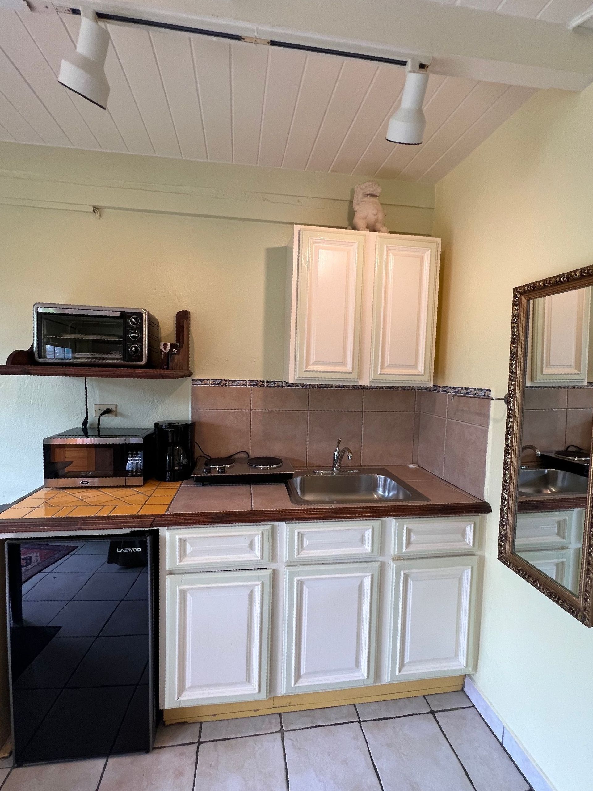 Small kitchen with white cabinets, mini-fridge, and stainless steel sink; a mirror hangs on the right.
