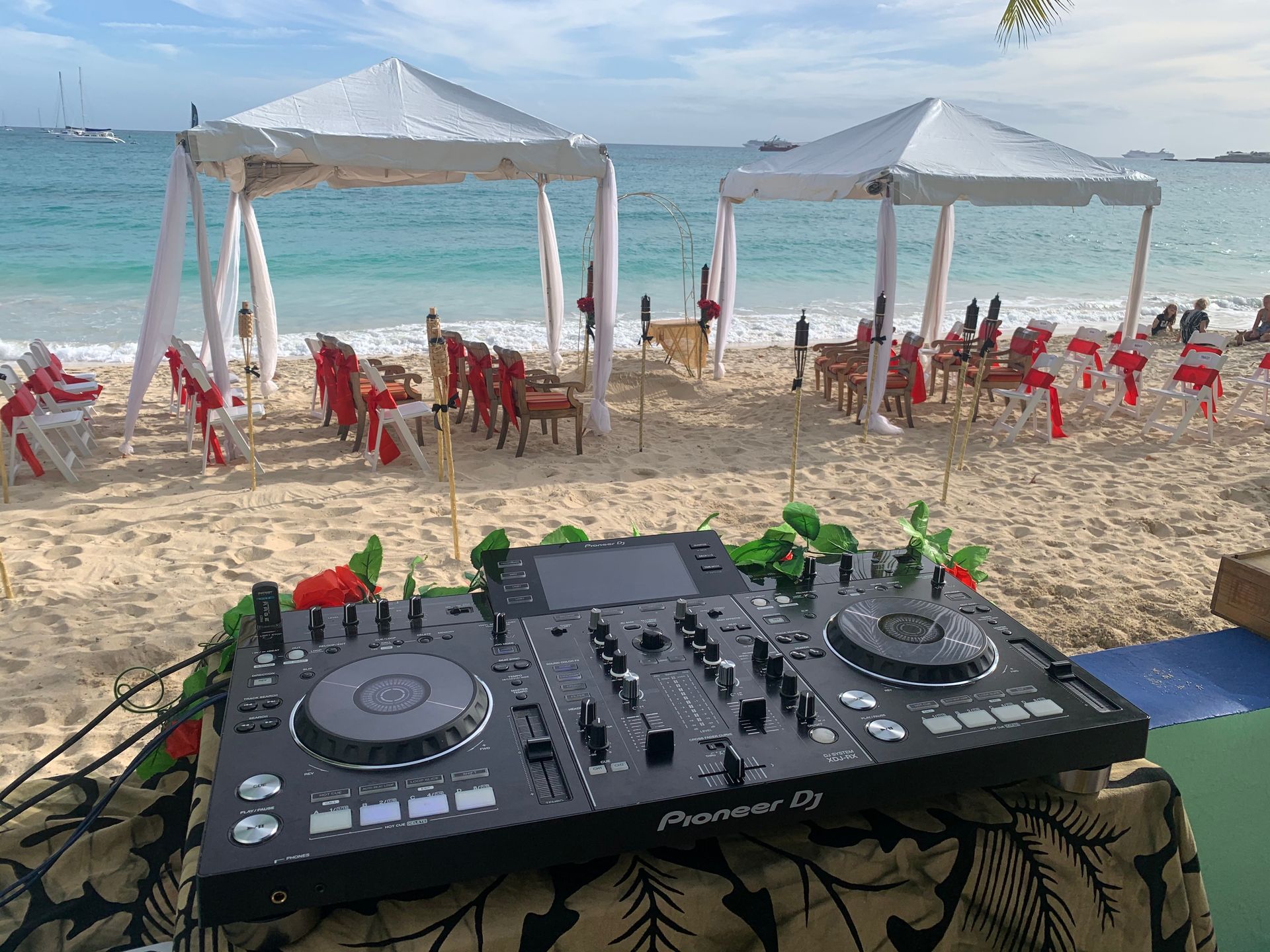 DJ setup on beach with white tents and wedding decor. Ocean and blue sky in background.
