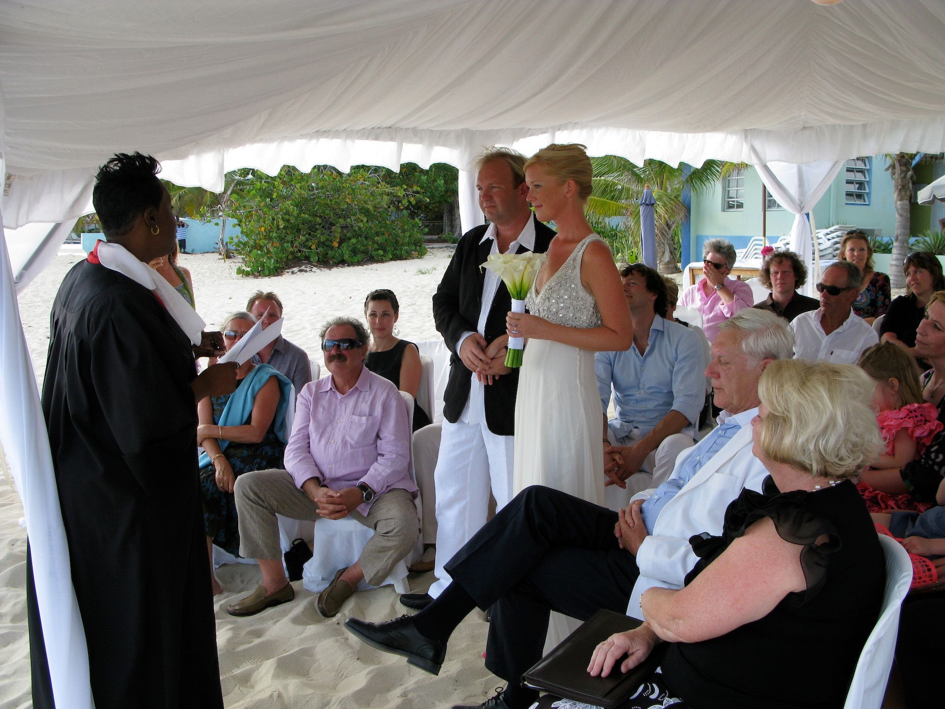 Wedding ceremony on a beach: Couple exchanging vows under a white tent, guests seated, officiant in black.