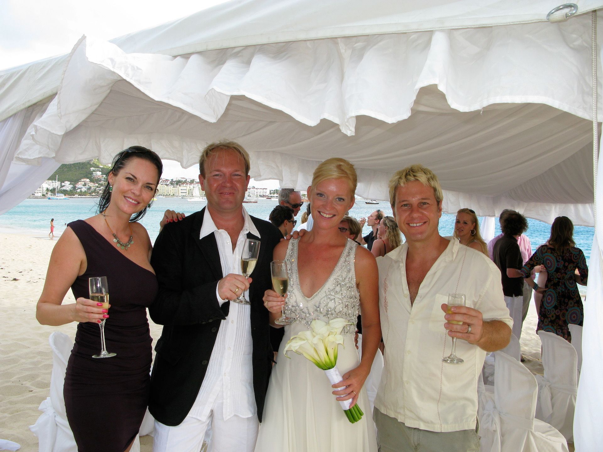 Wedding group on a beach, holding champagne. Bride in white dress, others in semi-formal attire under a tent.