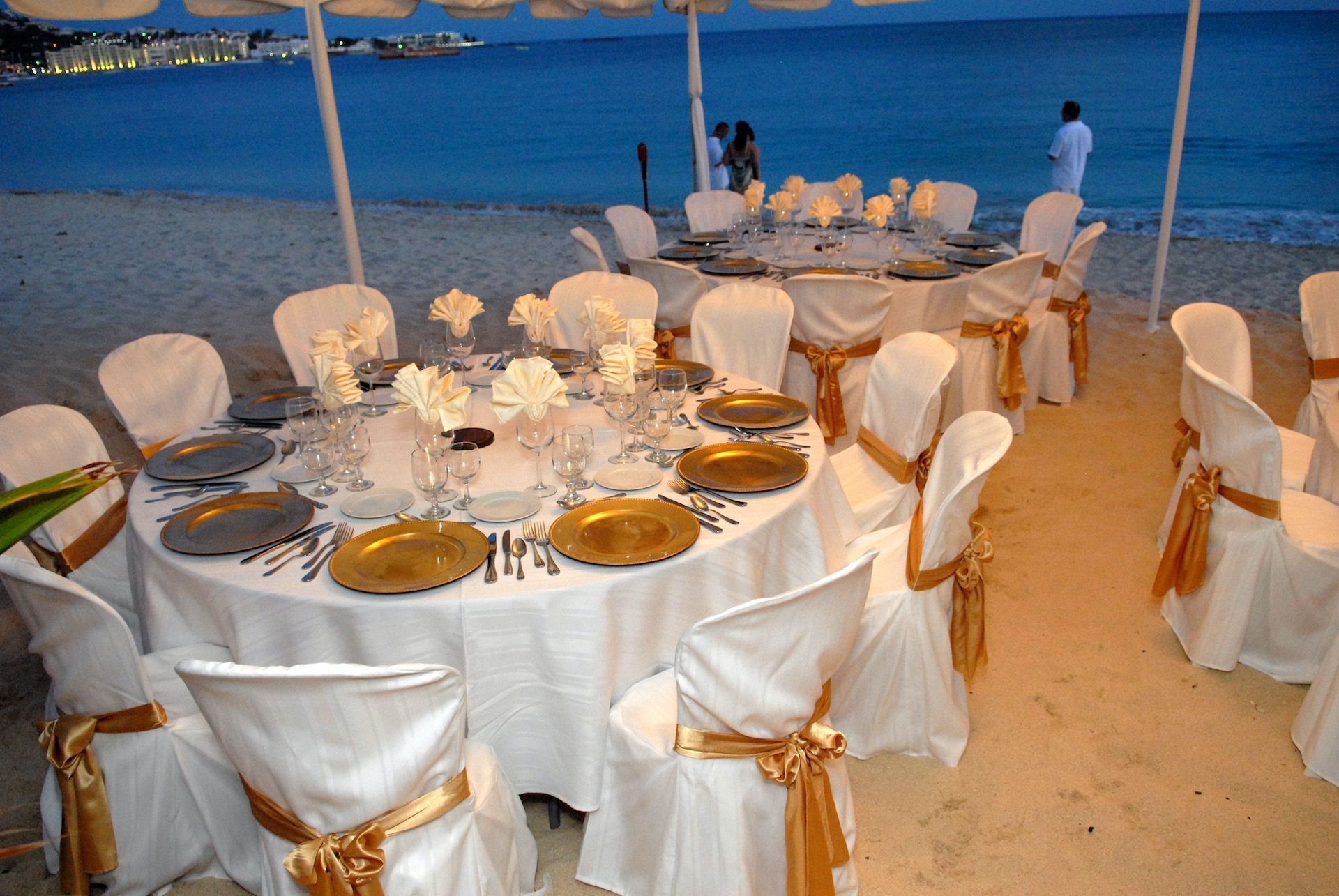 Beachfront wedding reception with round tables set for dinner. White chairs with gold sashes.