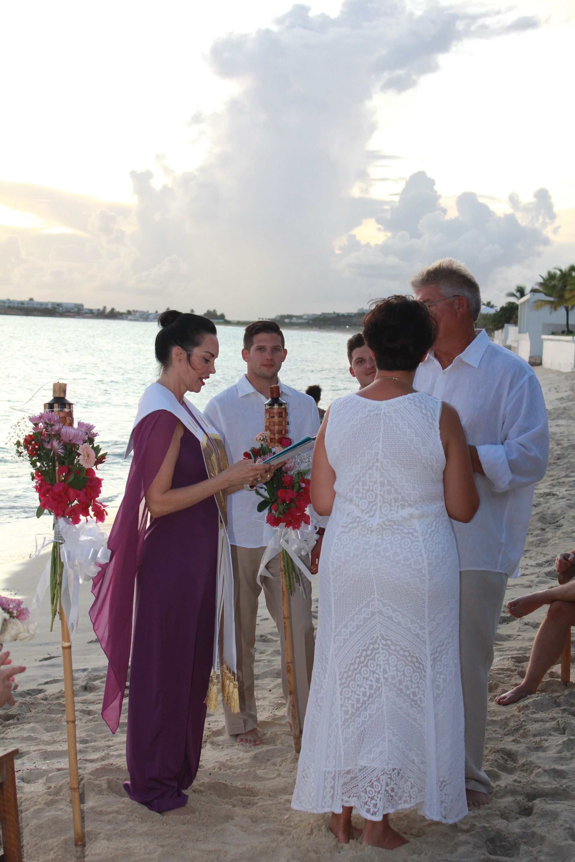 Wedding ceremony on a beach; couple, officiant, and family. Bride in purple, groom in white, guests on sand.